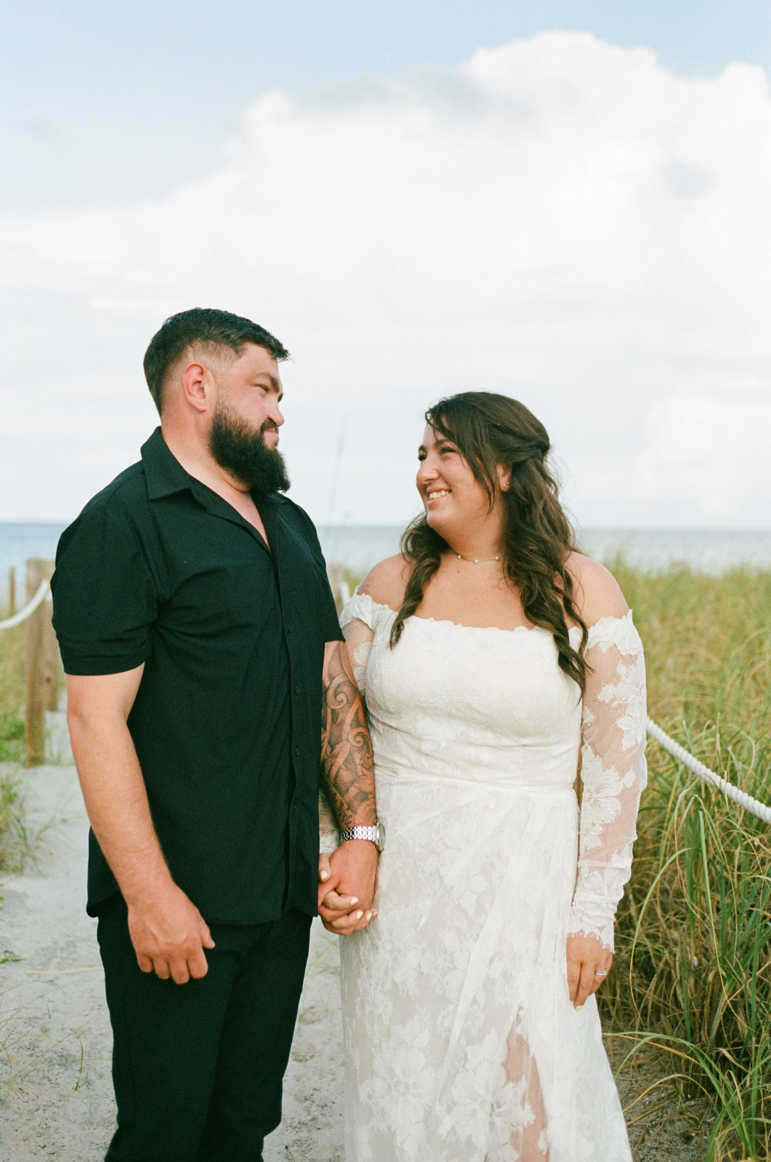 A couple holding hands, standing on a sandy beach near the water, with a cloudy sky above, during daytime.