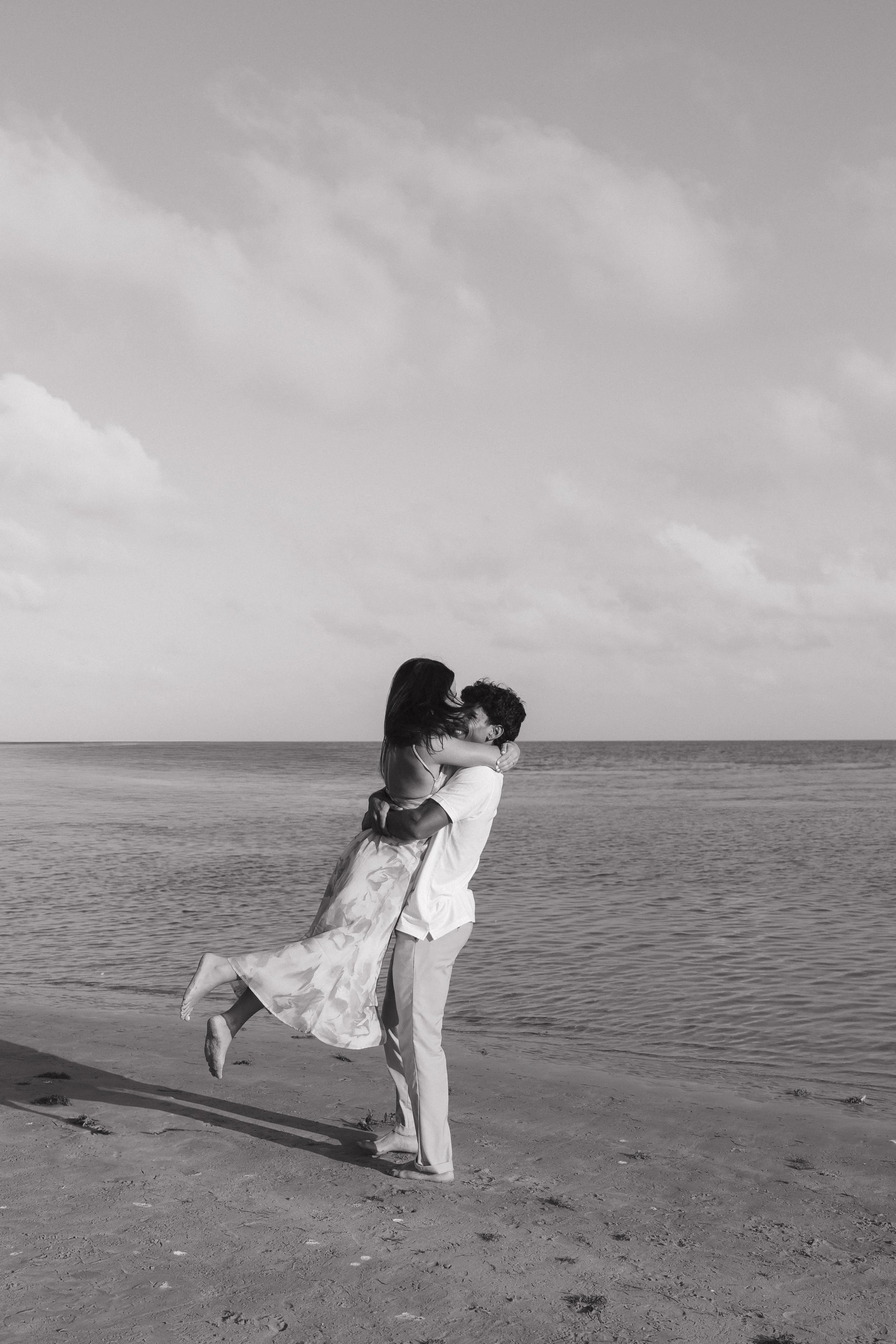 A couple on a beach, with the woman lifted in the air by the man, both smiling and embracing each other, with the ocean and cloudy sky in the background.