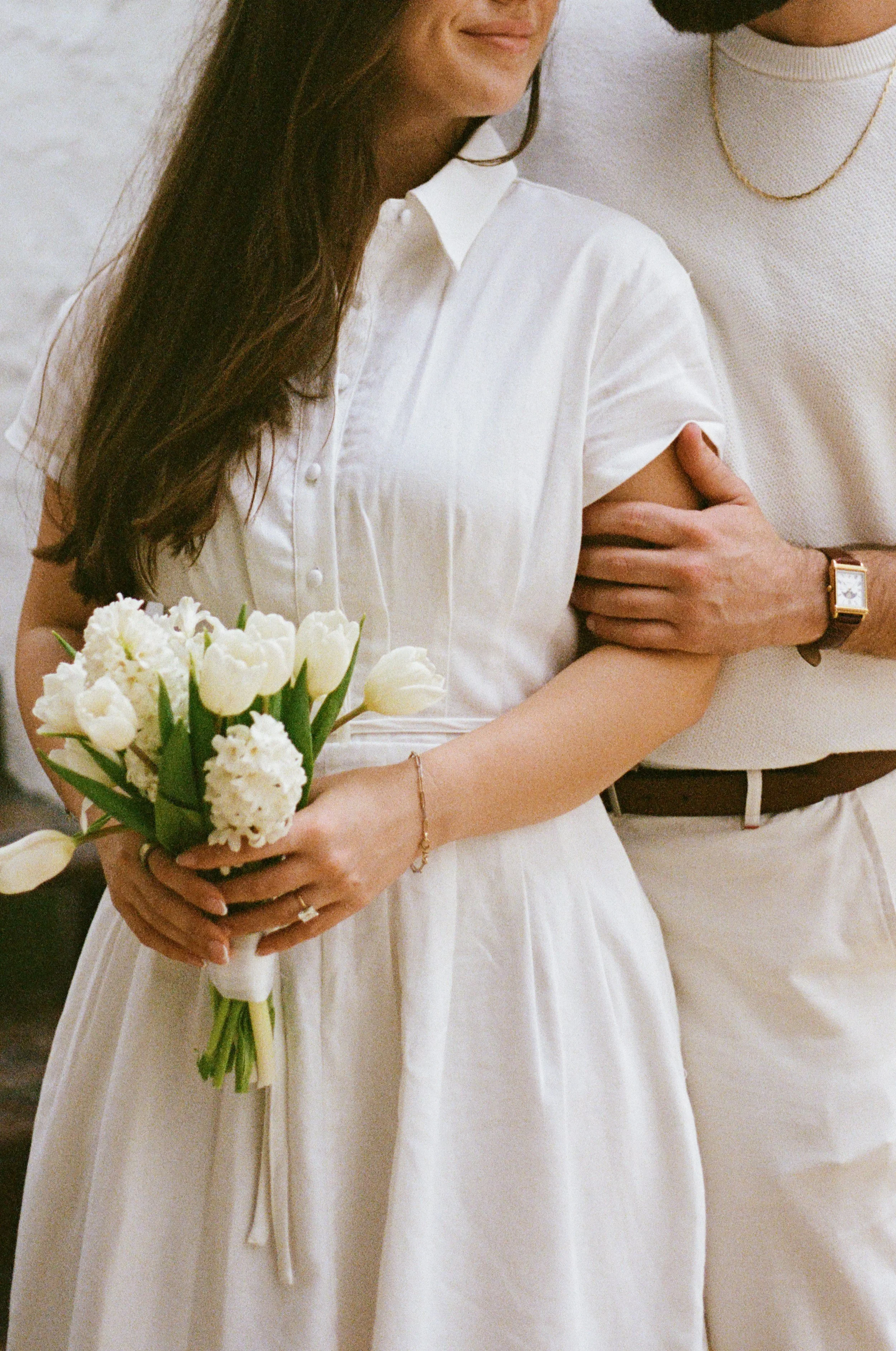 A woman in a white dress holding a bouquet of white flowers, standing next to a man wearing a white shirt and a gold watch, with their arms intertwined.