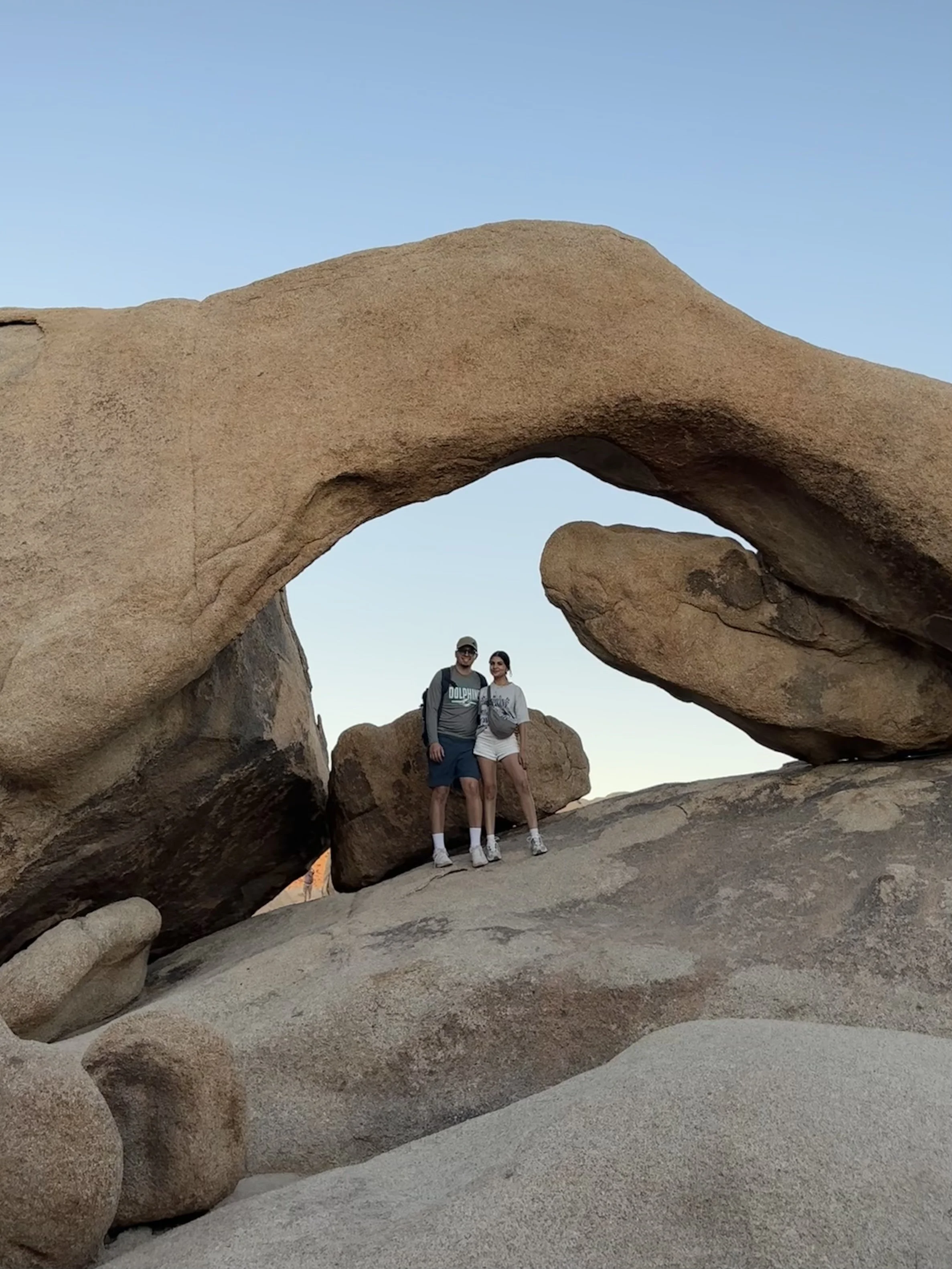 Two people standing underneath a large natural rock arch formation in a desert landscape with a clear blue sky.