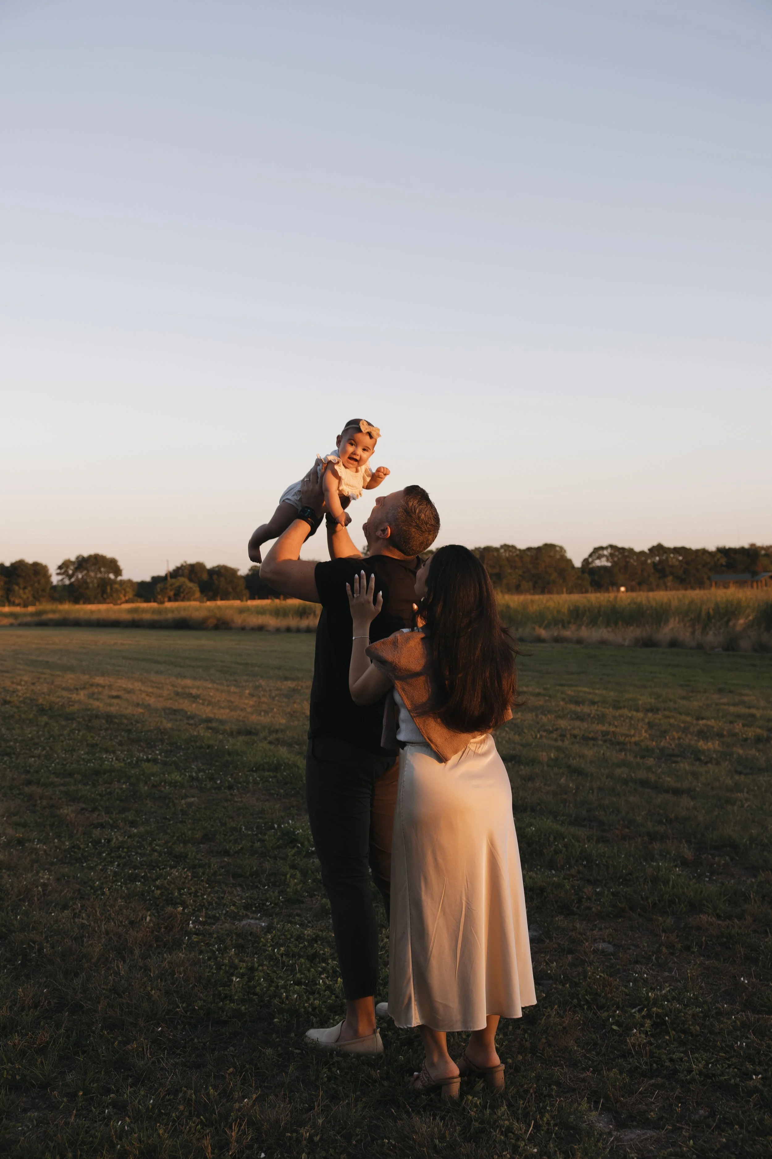 A family of three outdoors during sunset; a man lifts a baby girl in the air, woman stands nearby with her hands on the man's waist, and trees and a field are in the background.
