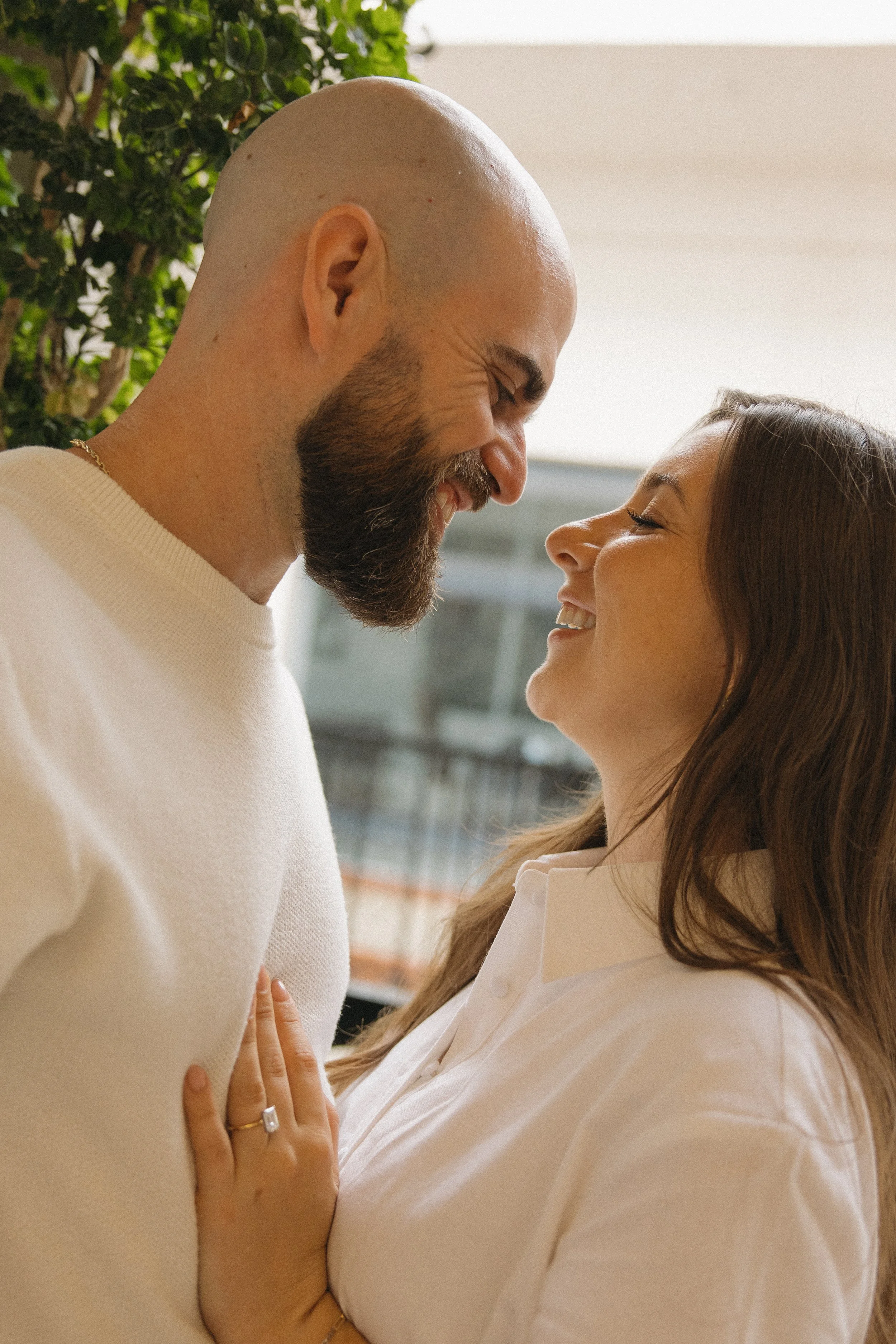 A man and woman sharing an intimate moment, touching foreheads and smiling at each other indoors, with greenery and a building in the background.