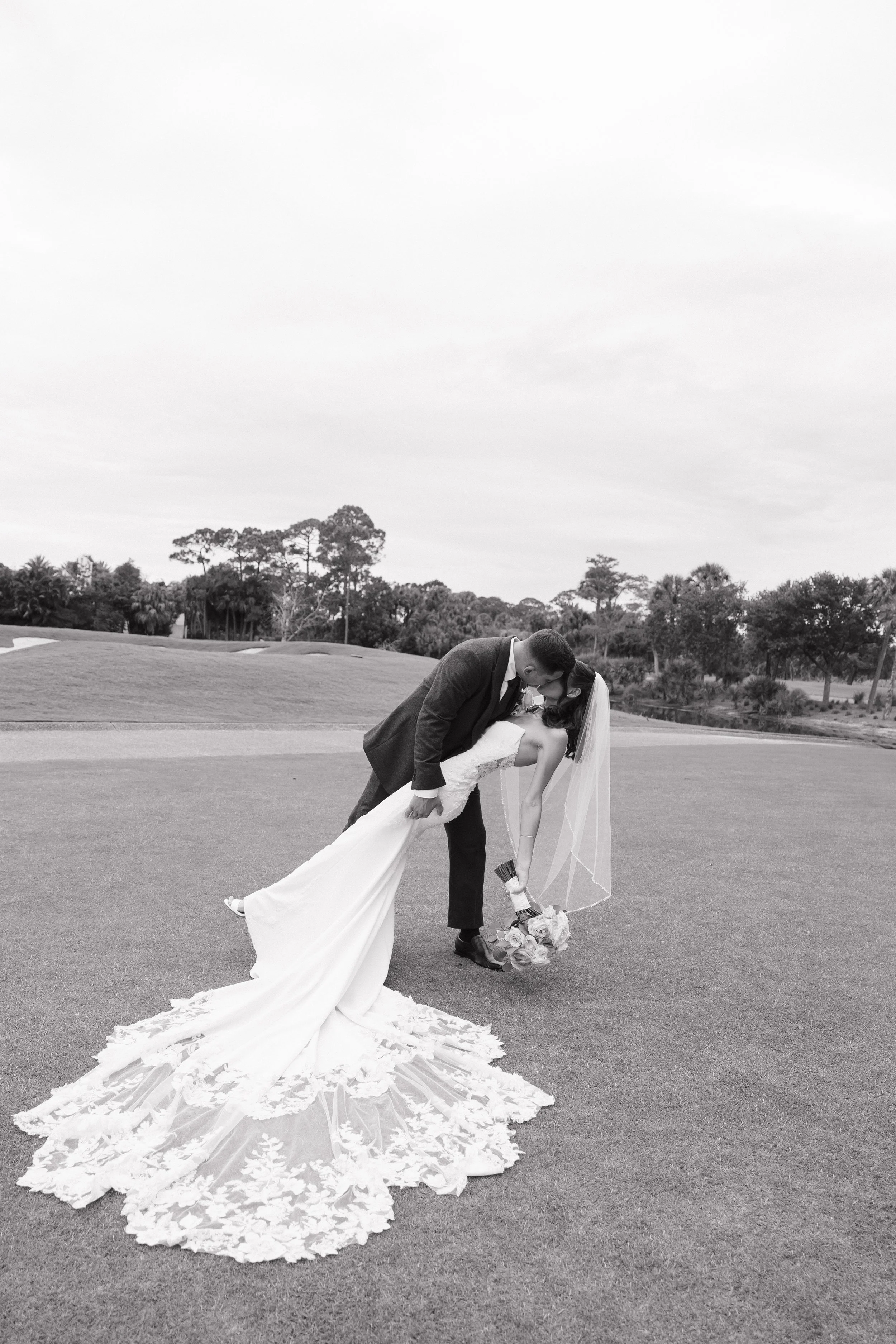 A bride and groom dancing on a grassy field with trees in the background. The groom is dipping the bride, who is holding a bouquet, and they are sharing a kiss.