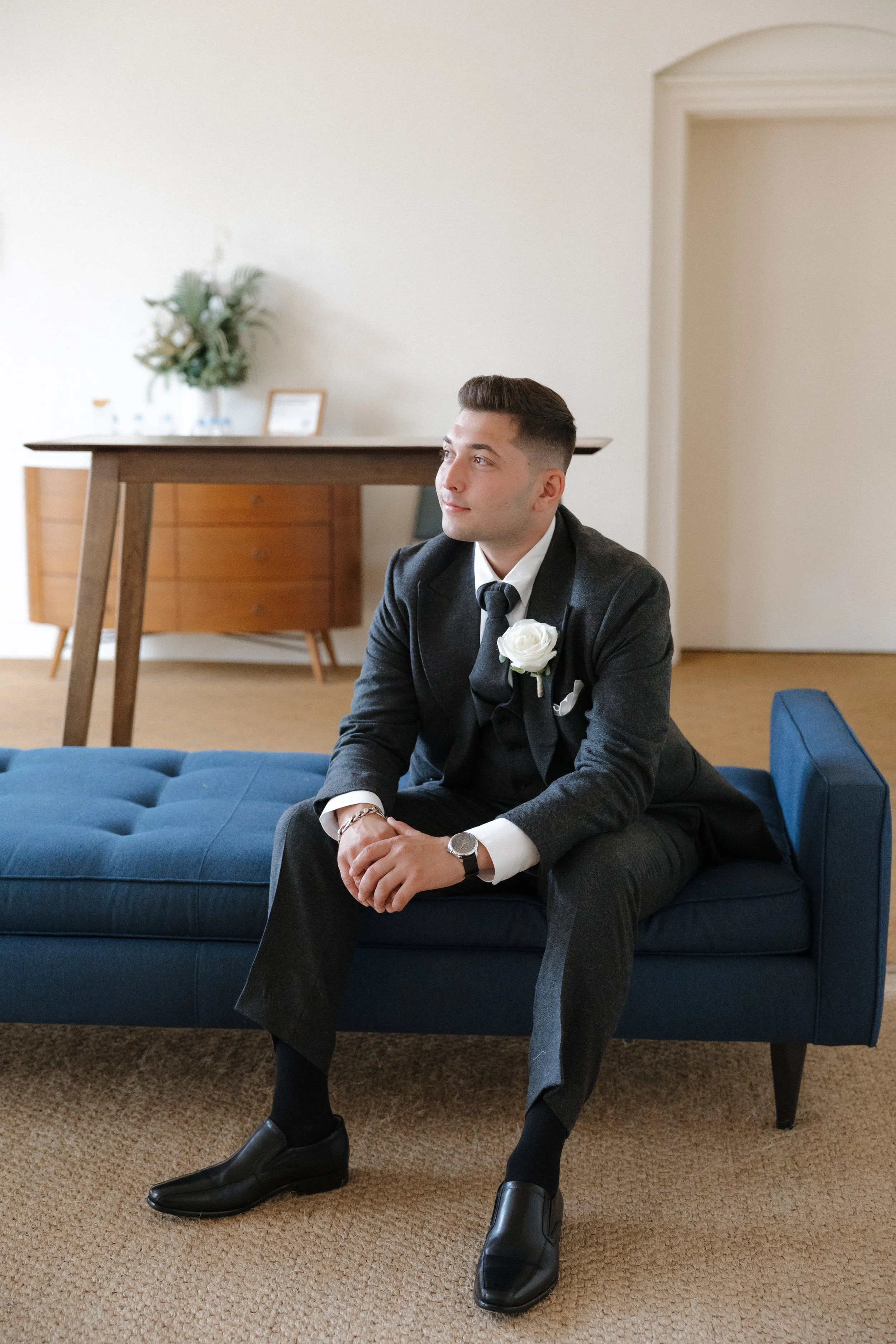A young man in a black suit with a white shirt and a black tie, wearing a white boutonniere, sitting on a blue upholstered bench in a room with beige carpet, a wooden sideboard, and a small framed photo and decorative plant on top.