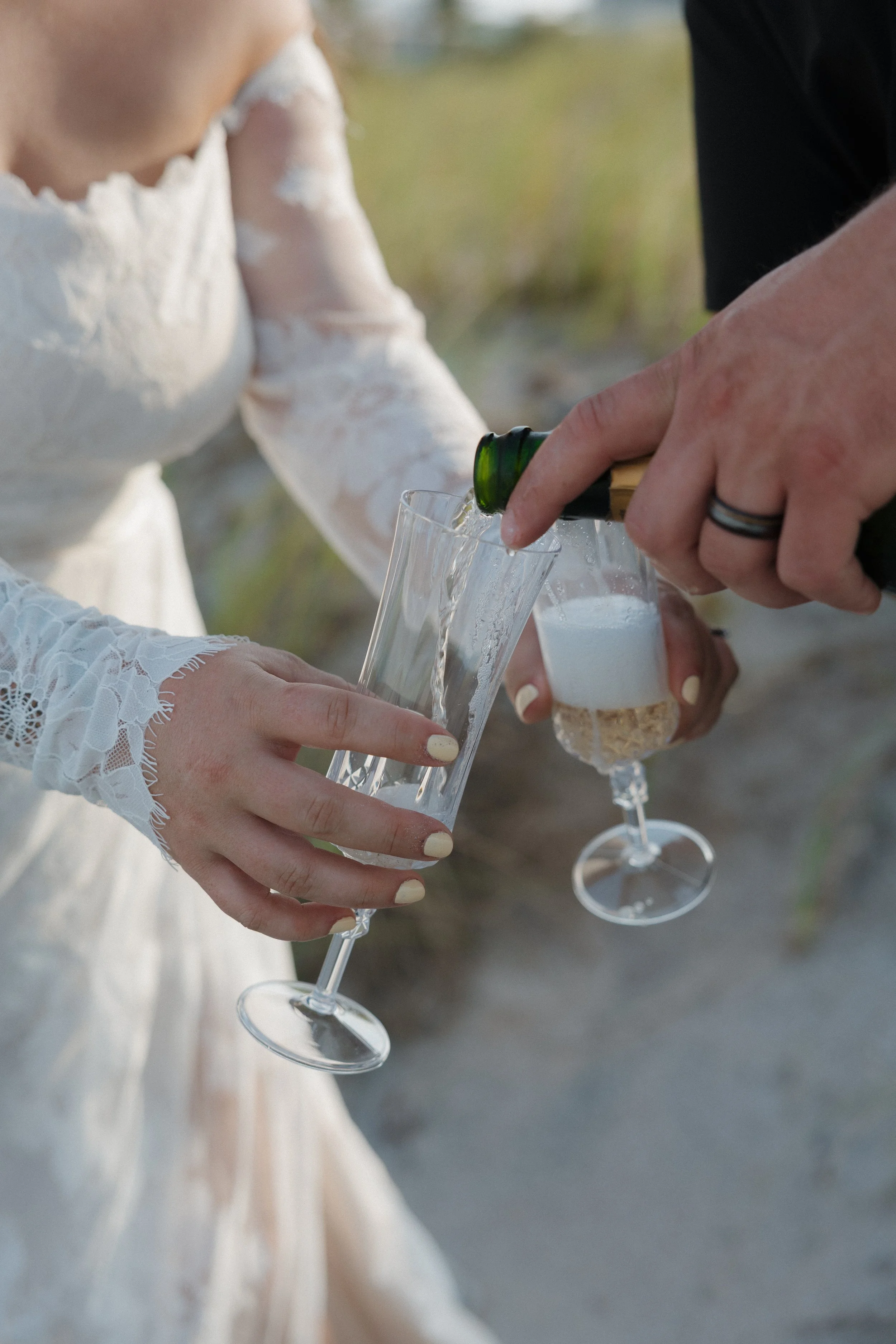 Someone in a white lace wedding dress pouring champagne into a flute glass held by another person, celebrating outdoors.