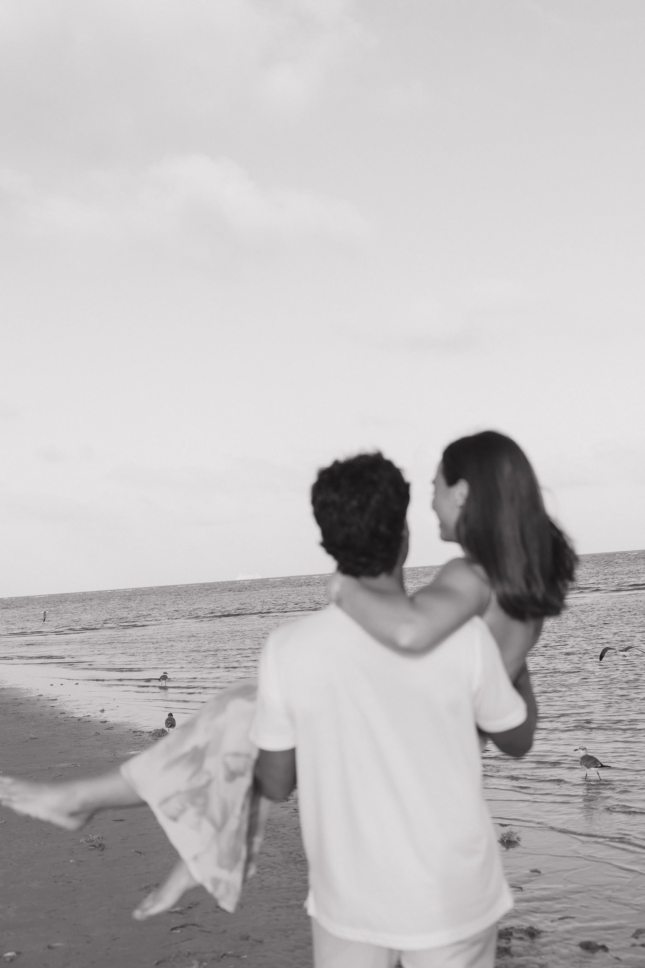 A couple embracing on the beach, looking at the ocean, with seagulls and people in the background.