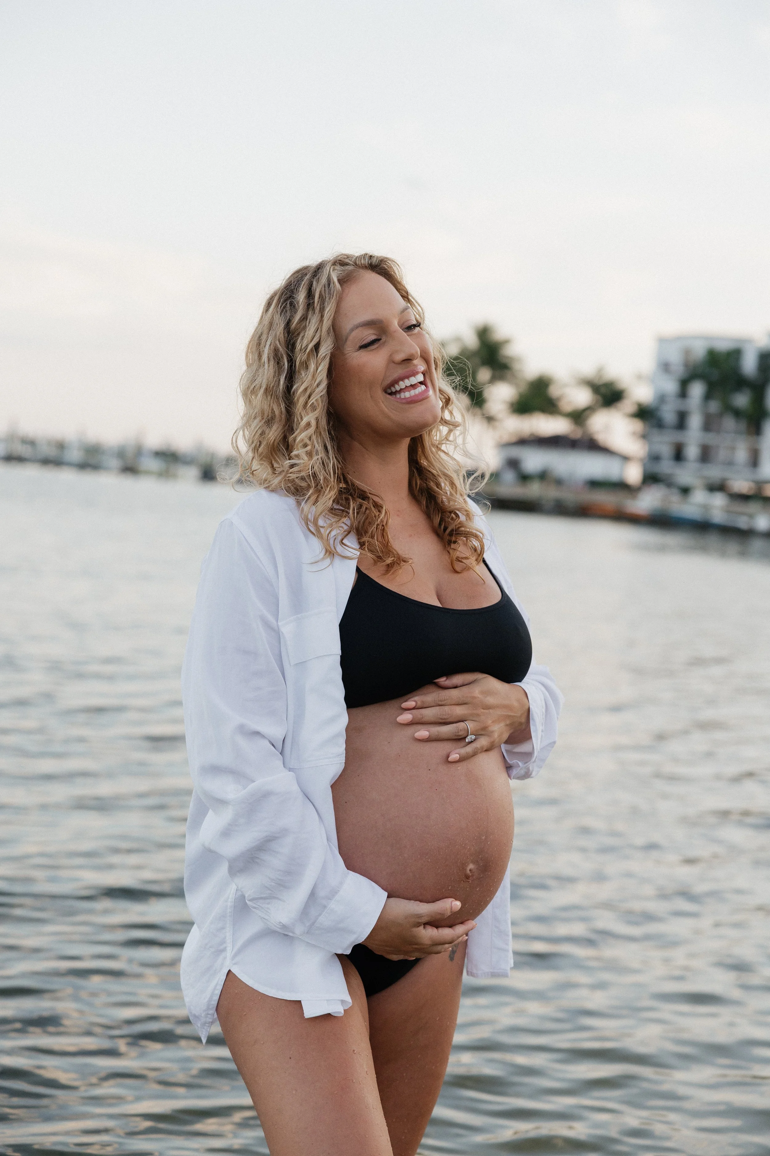 A pregnant woman with curly blonde hair smiling at the camera while standing by a body of water at sunset, wearing a black top, white shirt, and ring, with buildings and palm trees in the background.