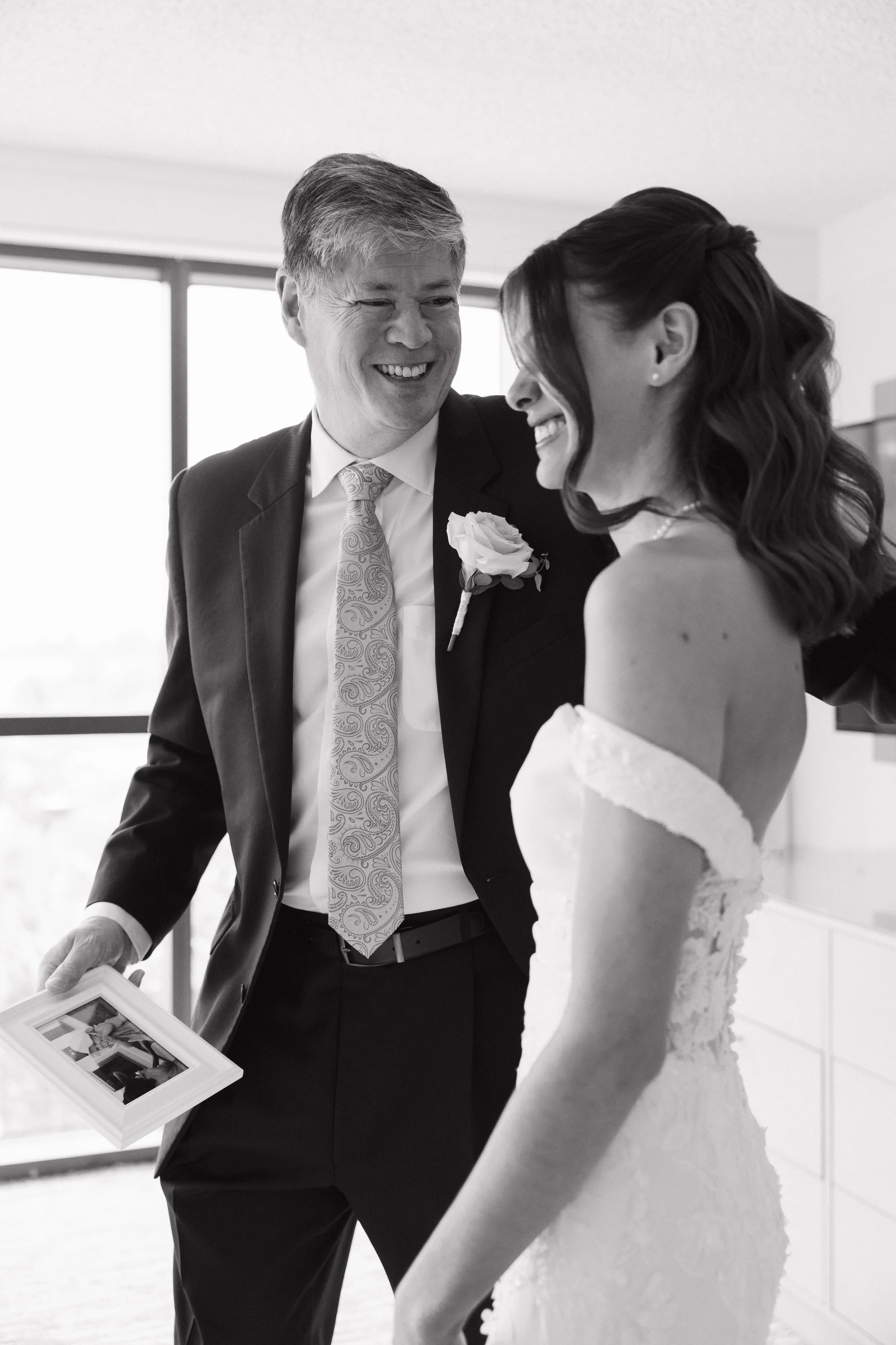 A black-and-white photo of a smiling man in a suit and a woman in a wedding dress looking at each other and laughing, possibly at a wedding or special event.