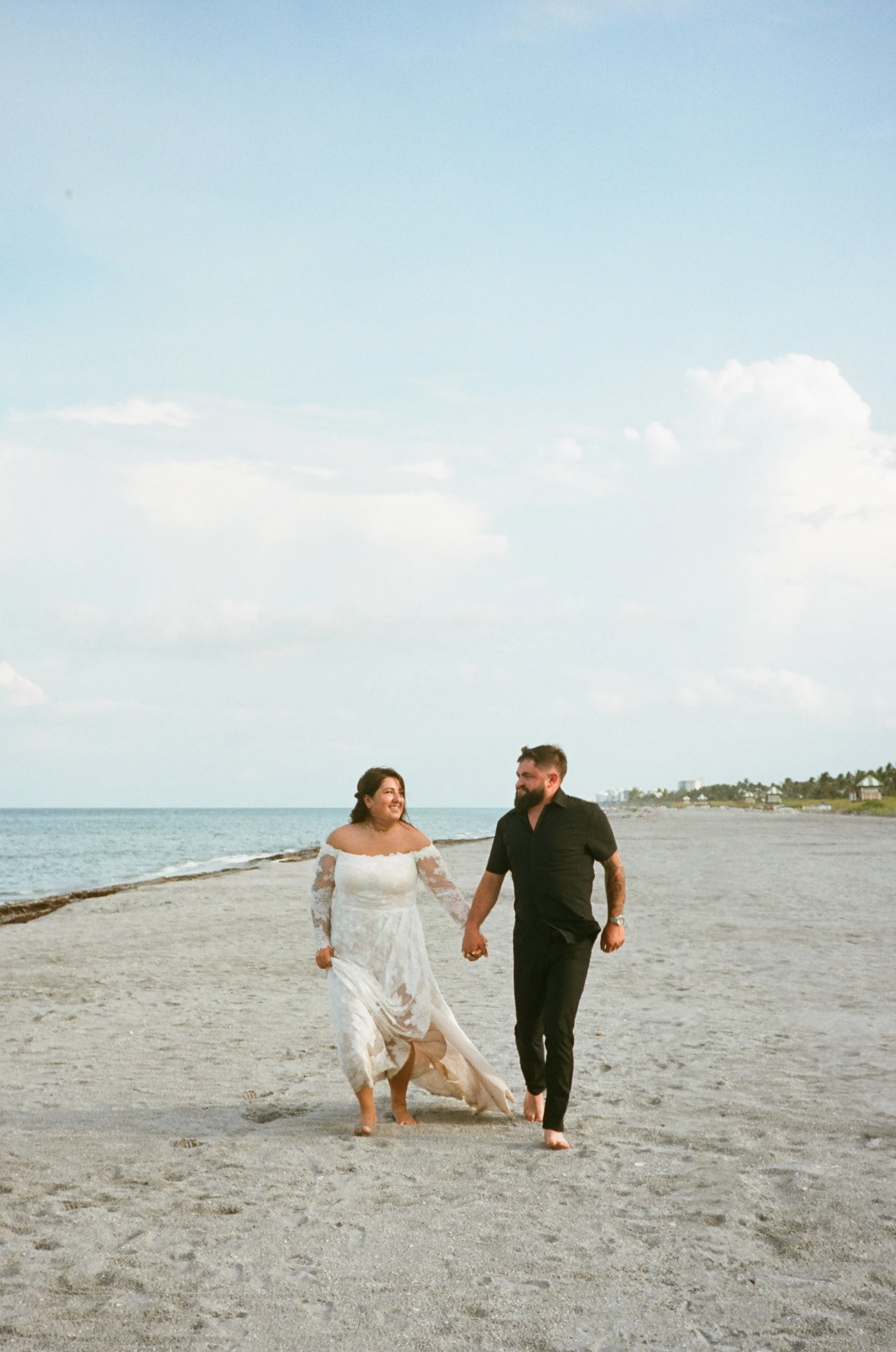 A bride and groom walking hand in hand on a beach, both smiling, with a cloudy sky and distant shoreline in the background.