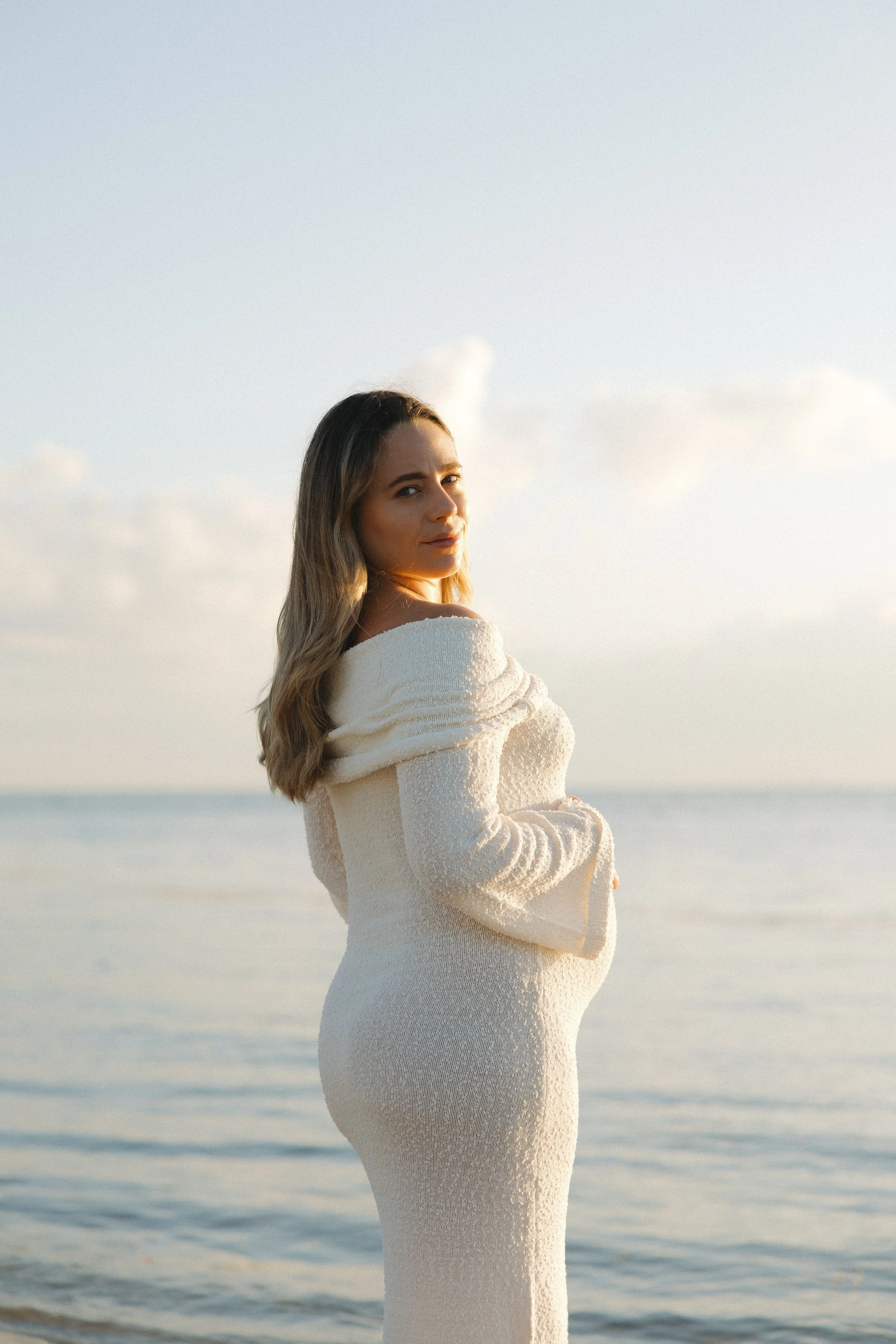 A pregnant woman standing at the beach during sunset, wearing a white knitted dress, with the ocean in the background.