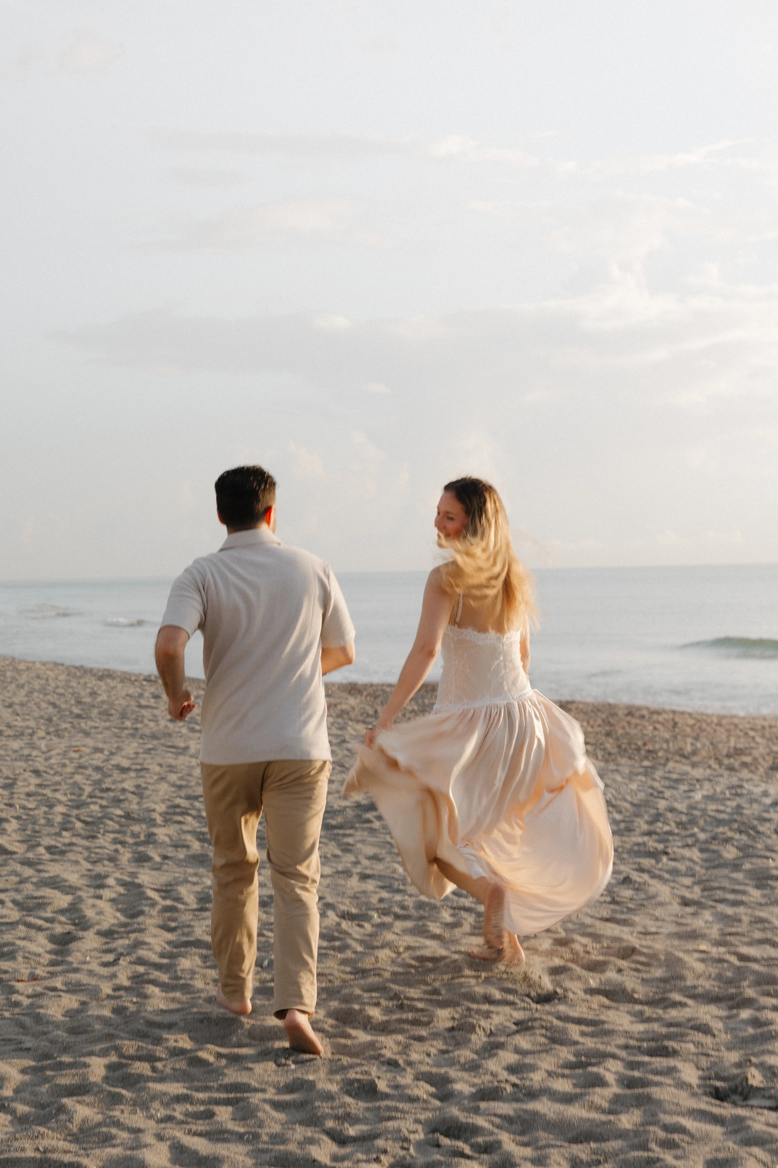 A man and woman walking barefoot on a sandy beach during sunset, with the ocean and sky in the background. The woman is wearing a flowing dress and smiling at the man, who is dressed casually.