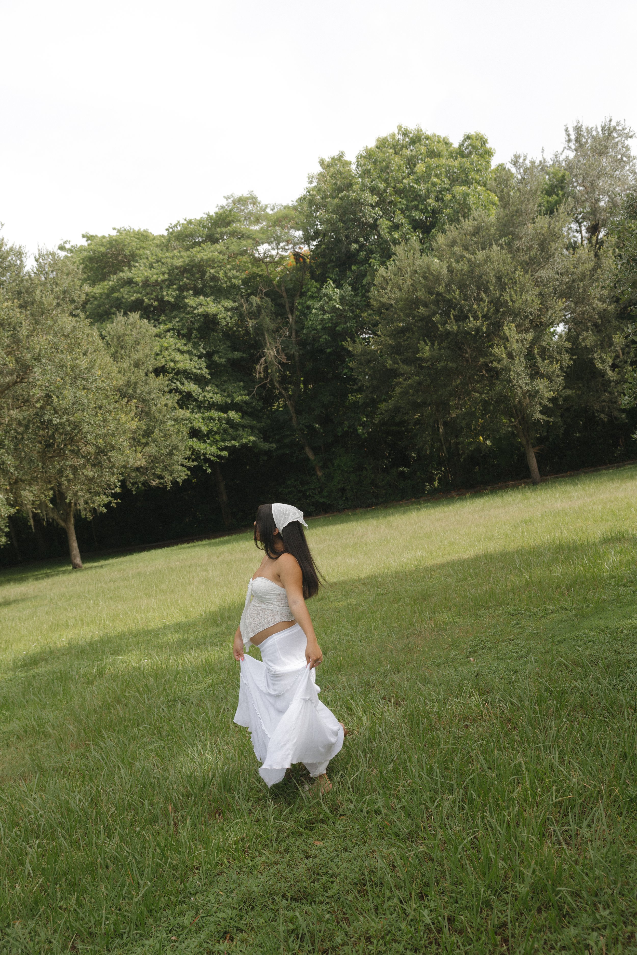 A woman in a white dress walking through a grassy field with trees in the background under a clear sky.