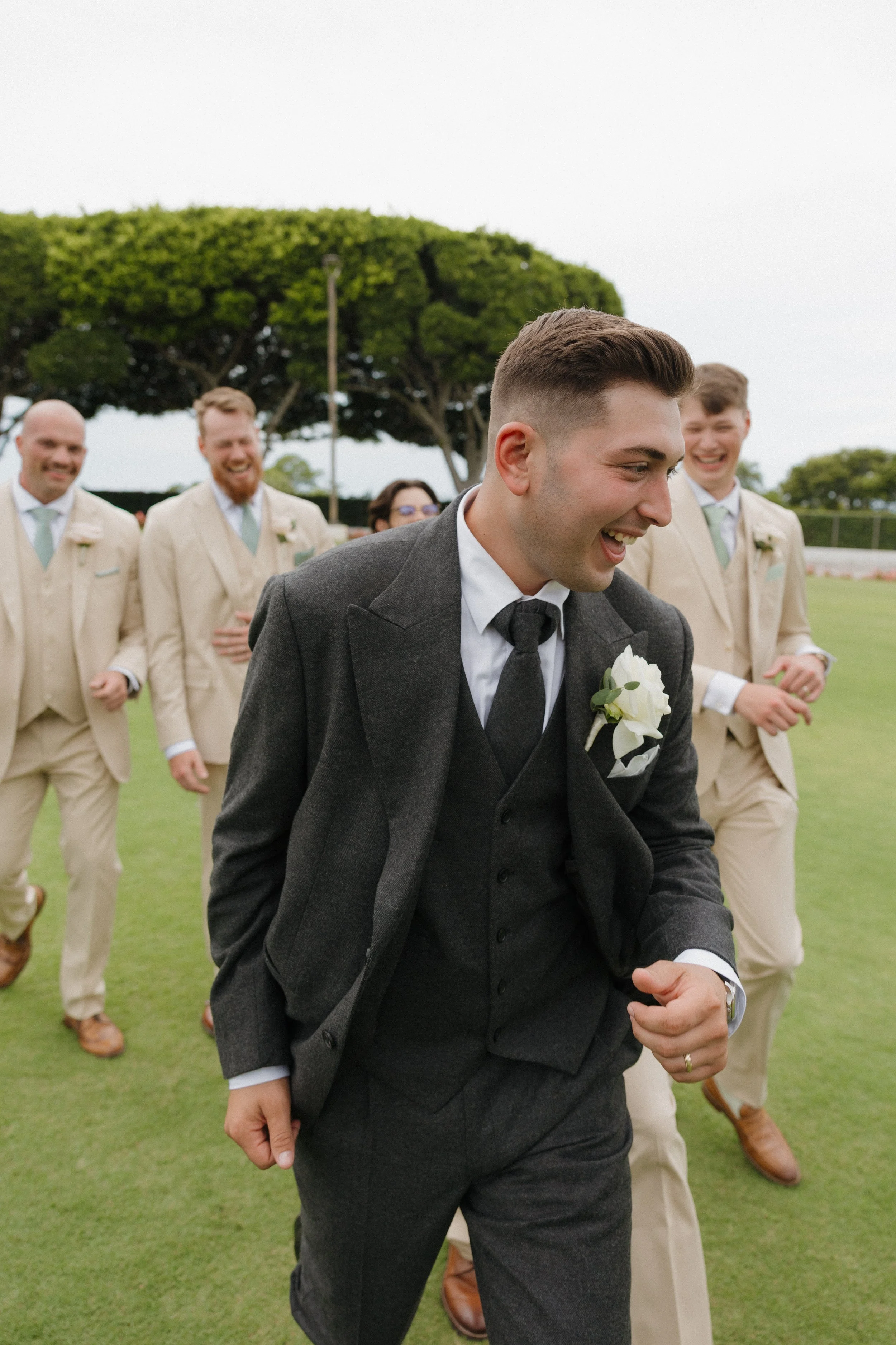 A group of men dressed in suits and ties, celebrating outdoors on a grassy field, with a large tree and cloudy sky in the background.