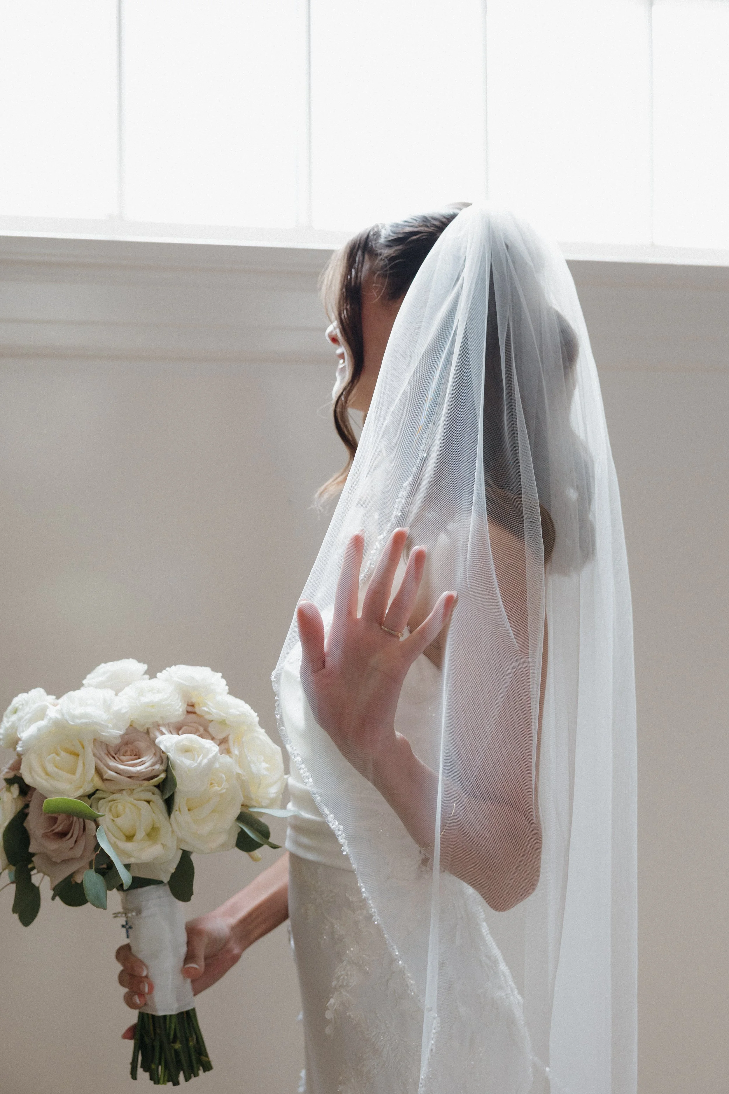 A bride with dark brown hair in loose waves, wearing a white wedding gown and veil, holding a bouquet of white and blush roses and greenery, standing near a window with bright natural light.
