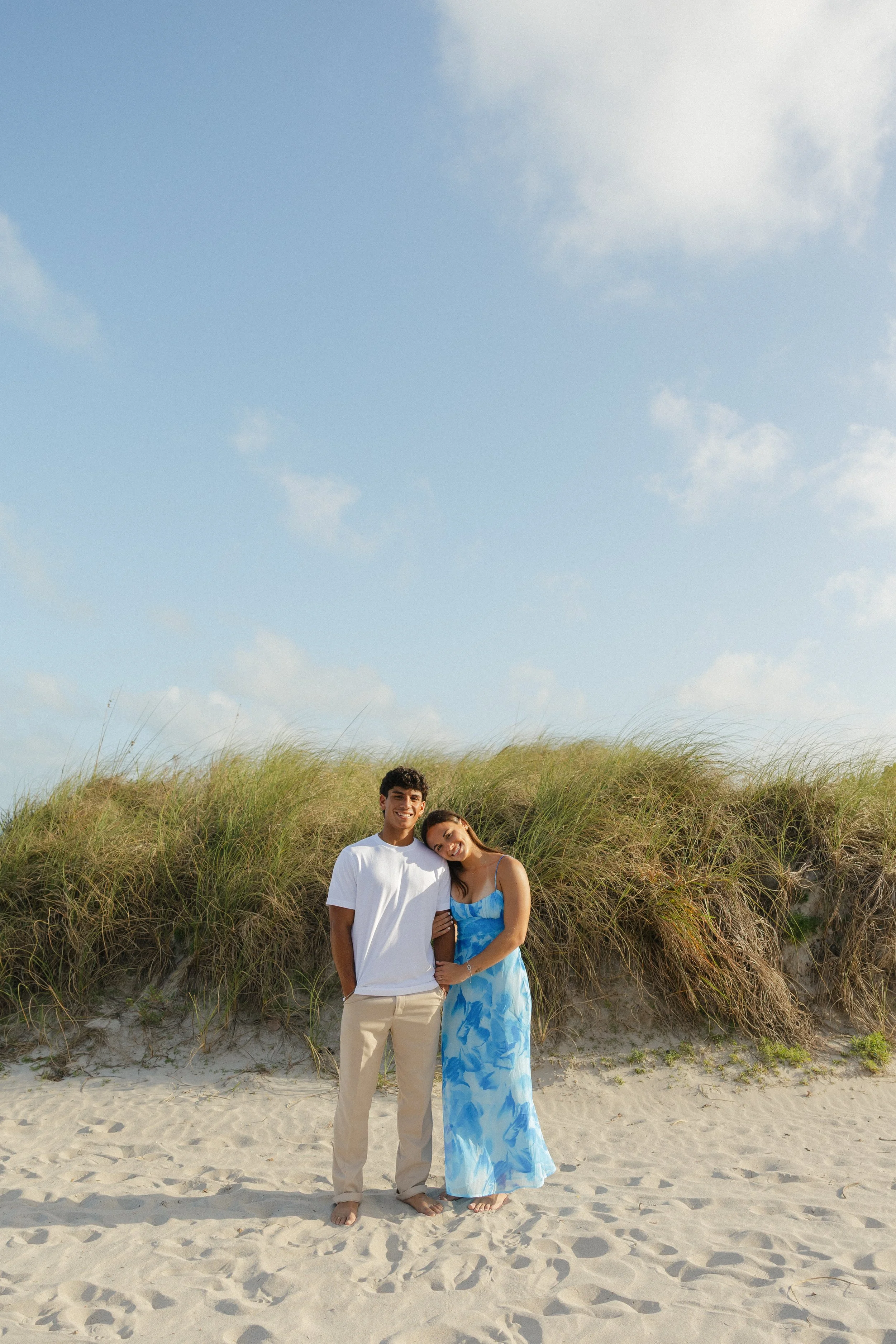 A smiling young man and woman standing together on a sandy beach, with green dune grass behind them and blue sky with clouds above.
