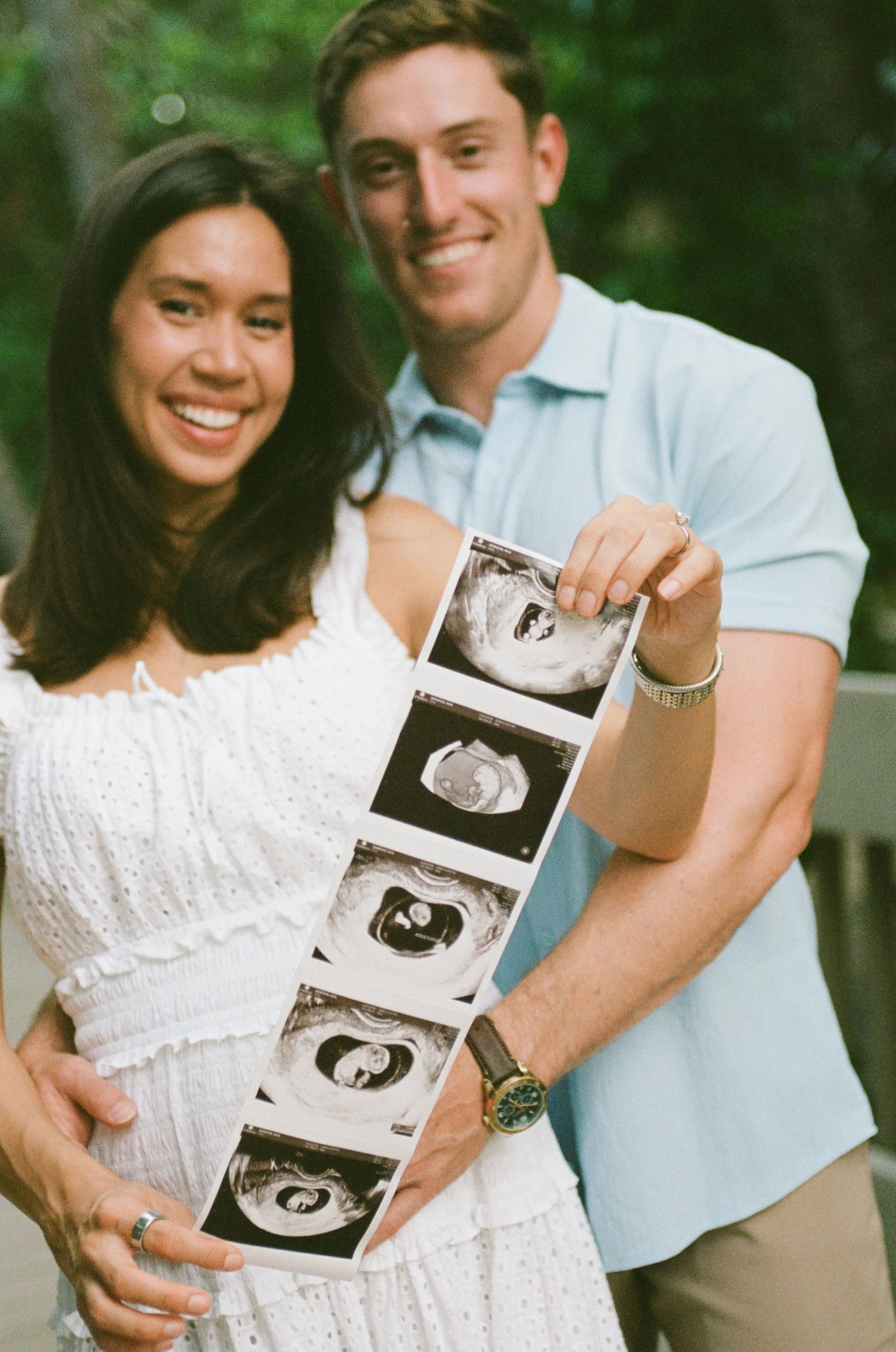 A couple stands outdoors, smiling as they hold ultrasound images showing a developing fetus.