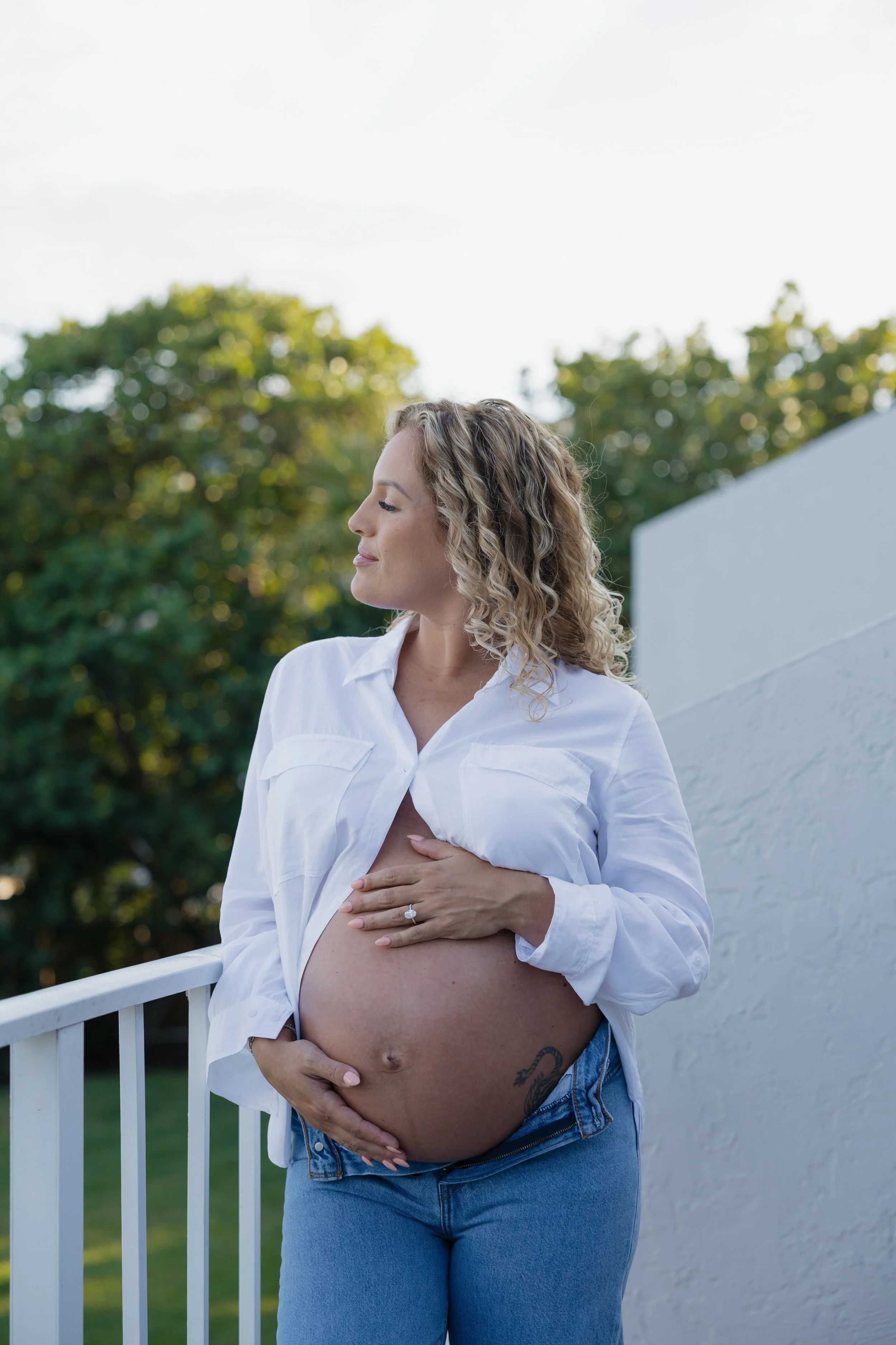 Pregnant woman standing outdoors on a balcony in front of a white wall, with her eyes closed and hand on her belly, wearing a white shirt and blue jeans.
