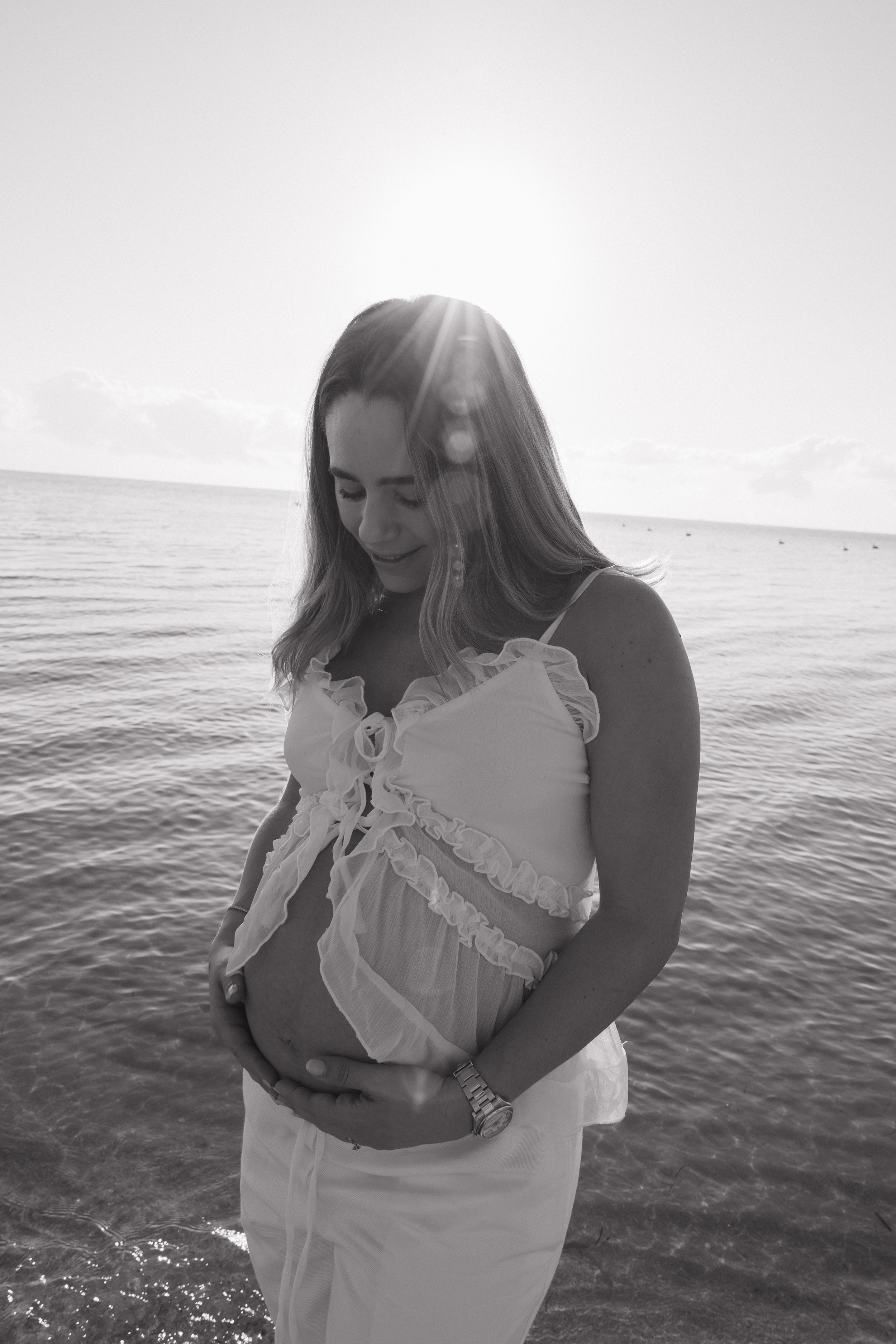 A pregnant woman standing in the shallow water at the beach, wearing a light-colored dress with ruffles, smiling gently and looking down at her belly, with the sun shining brightly in the sky behind her.