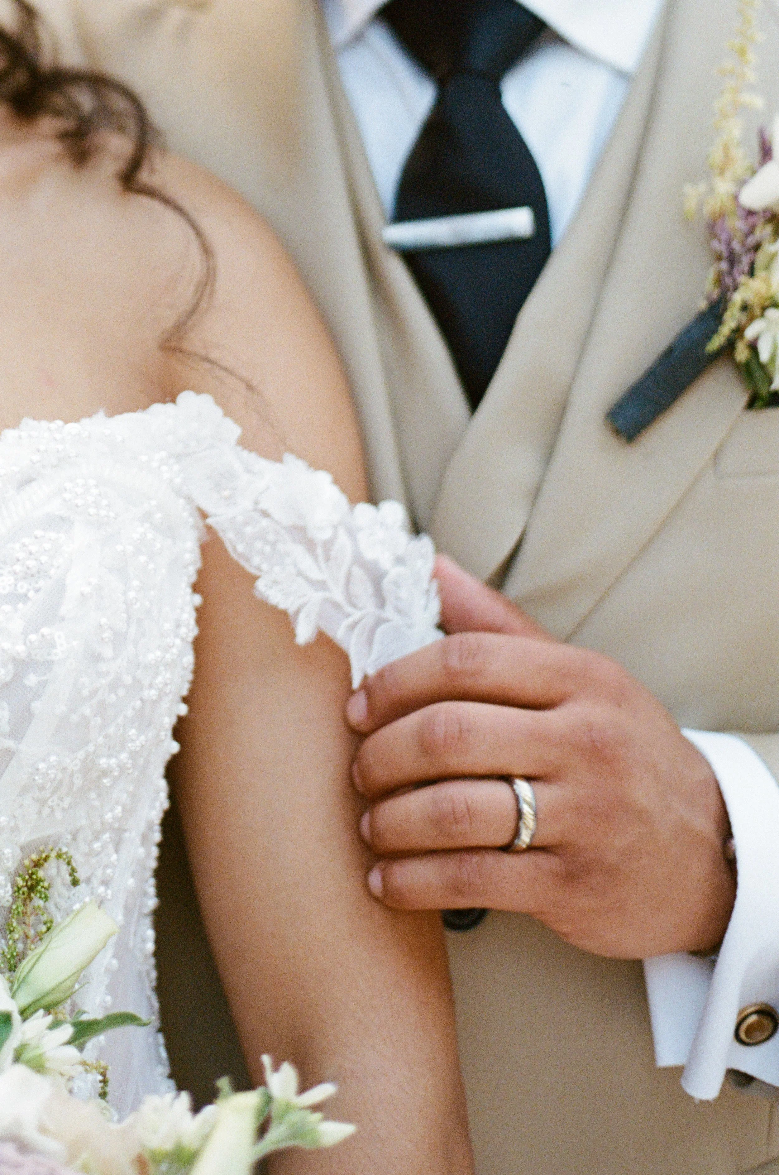Close-up of a groom holding a bride's arm during a wedding, with a mirror reflecting his tuxedo and a boutonniere on his suit.