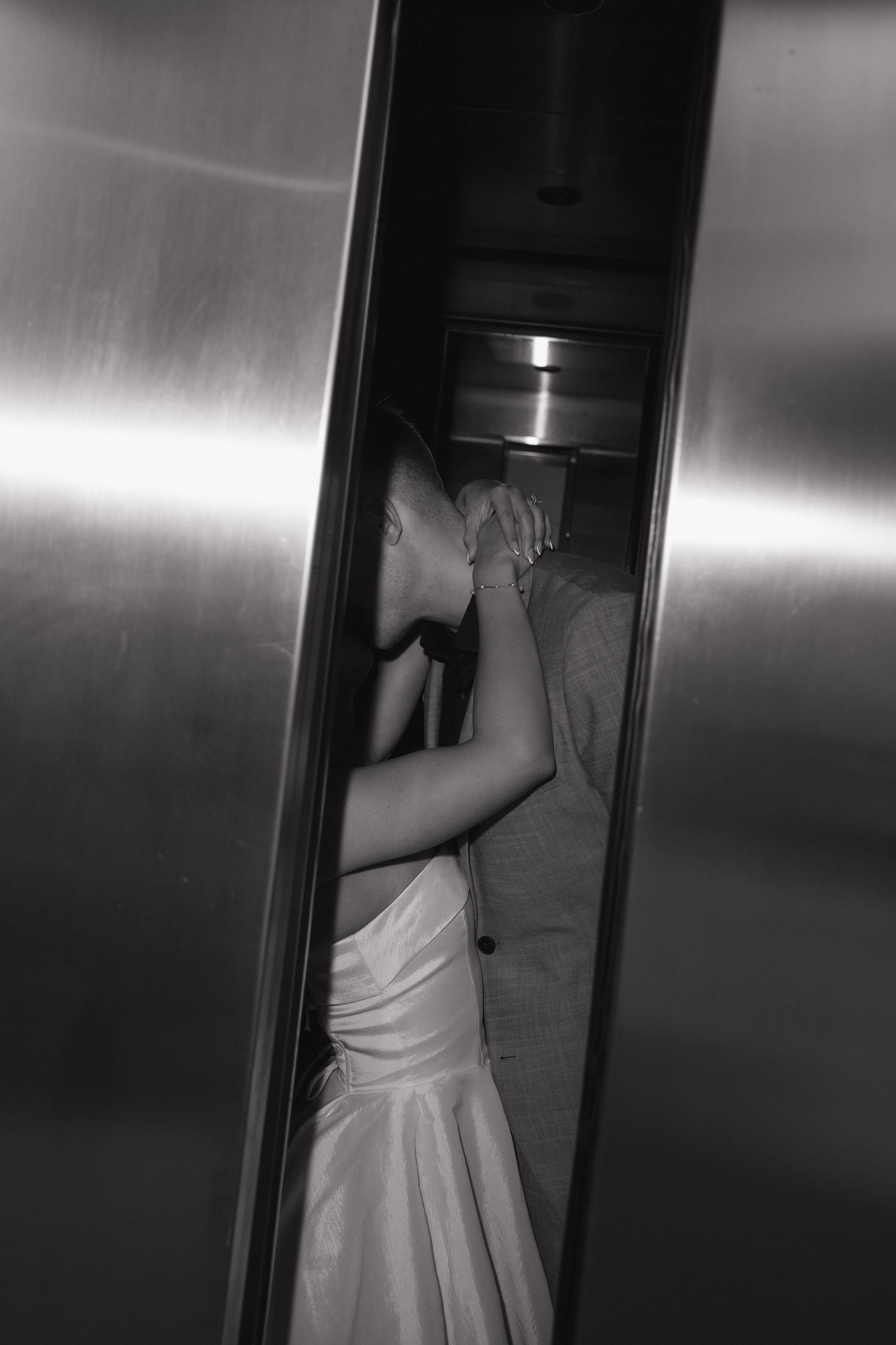 A woman in a wedding dress and a man in a suit taking a kiss inside an elevator, filmed through the elevator reflection.