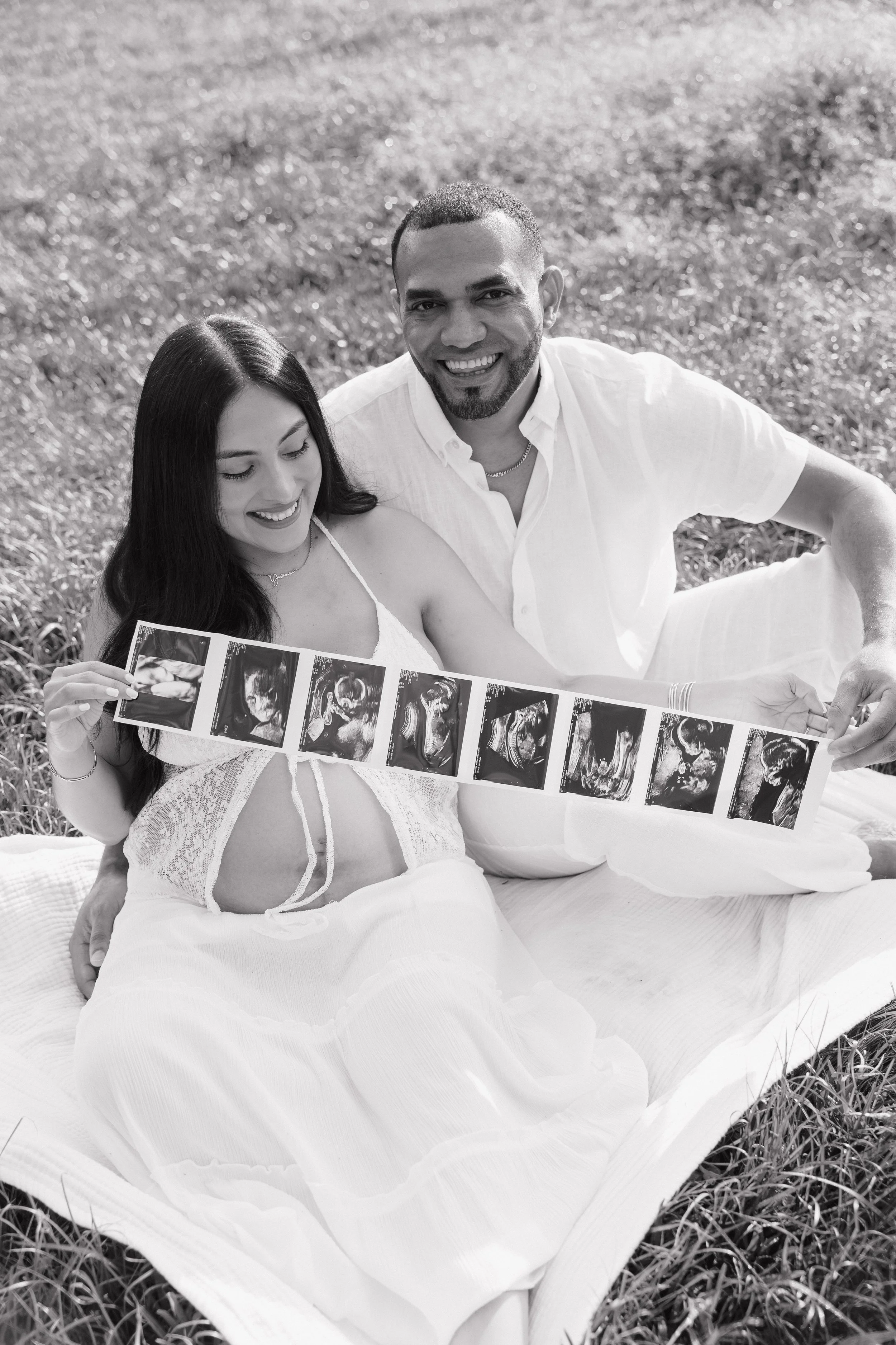 A smiling couple sitting on a blanket outdoors in a field, looking at ultrasound images.