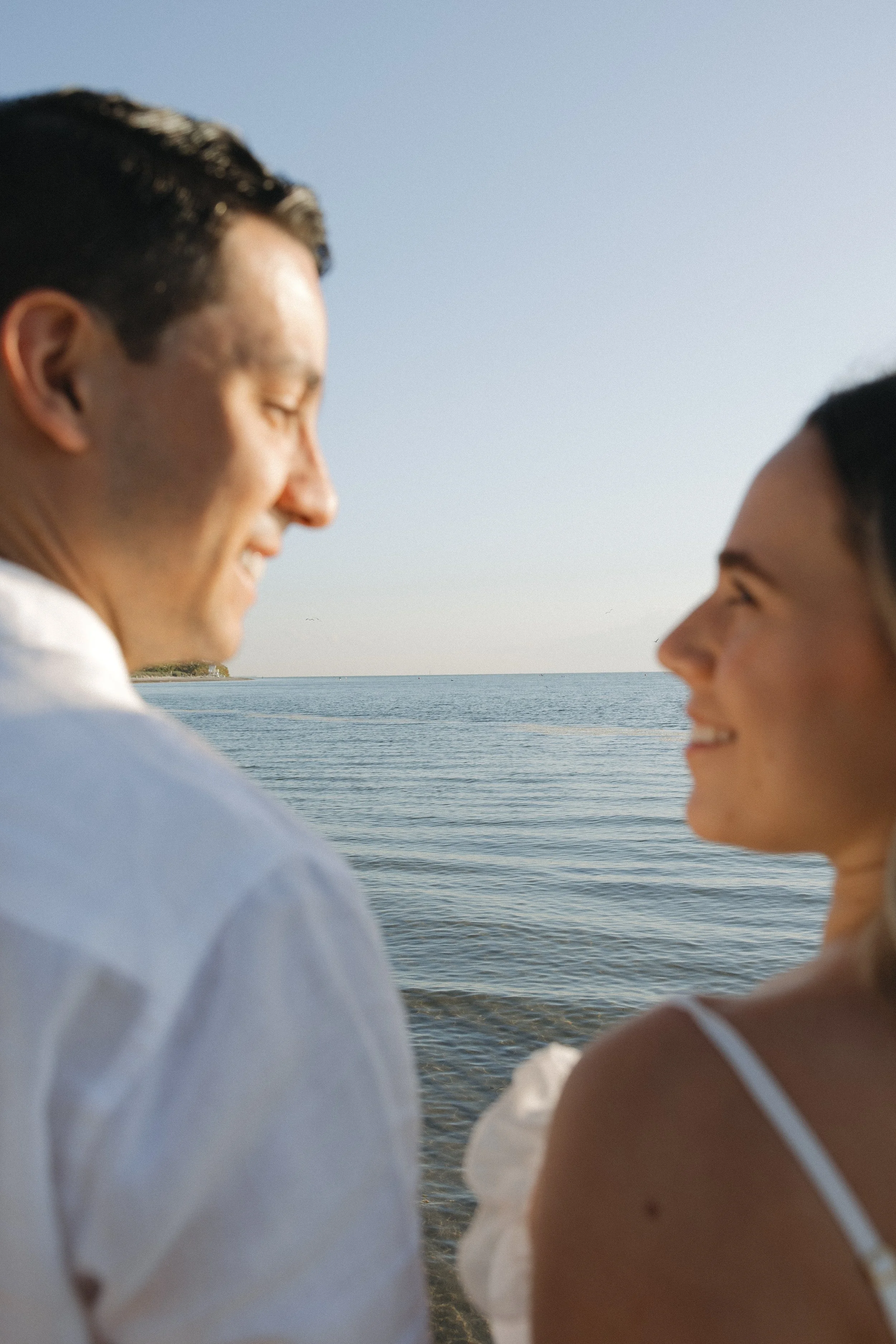 Close-up of a smiling man and woman facing each other at the beach, with water and sky in the background.