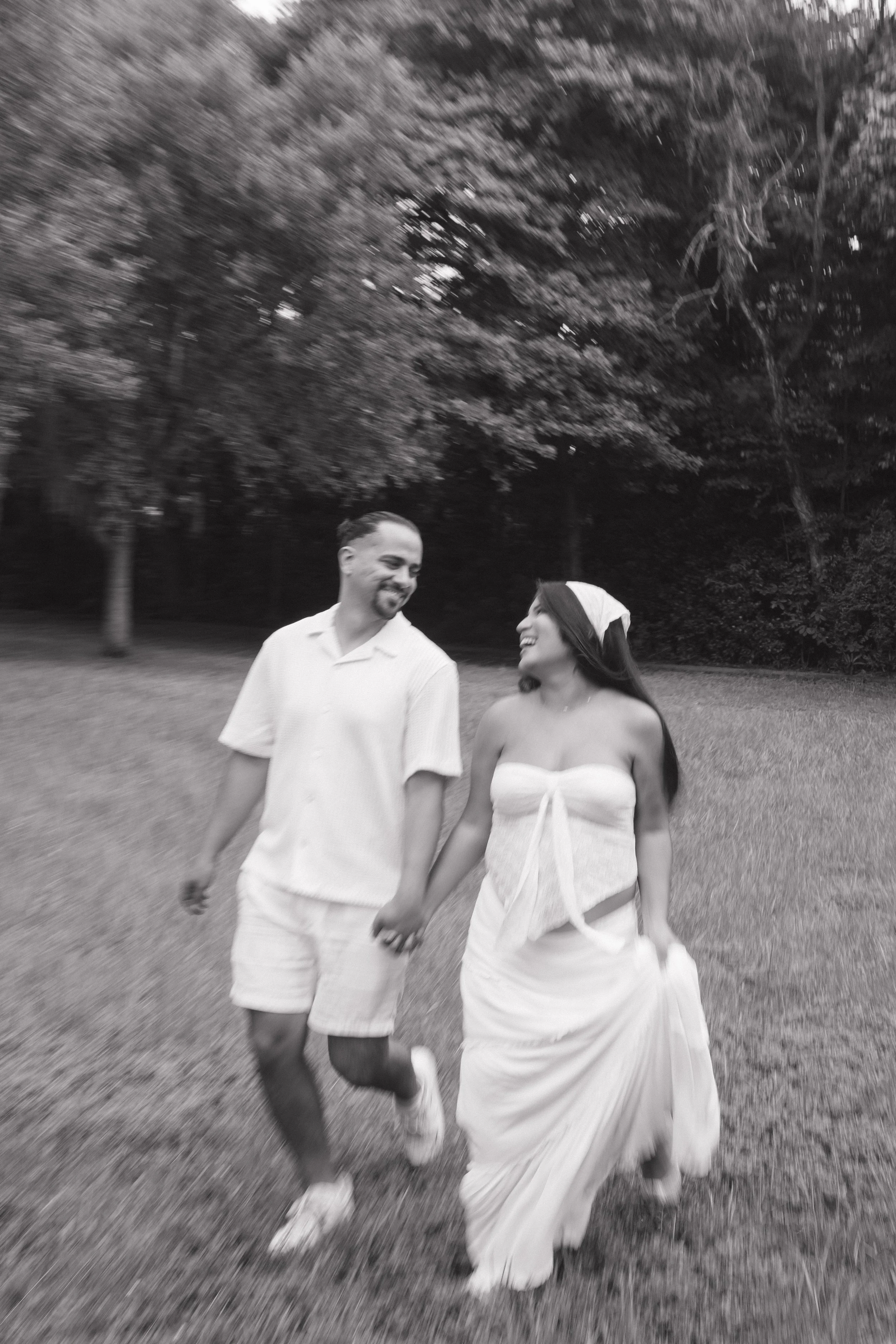 A black and white photo of a smiling couple walking hand in hand outdoors in a park or garden, surrounded by trees and grass.