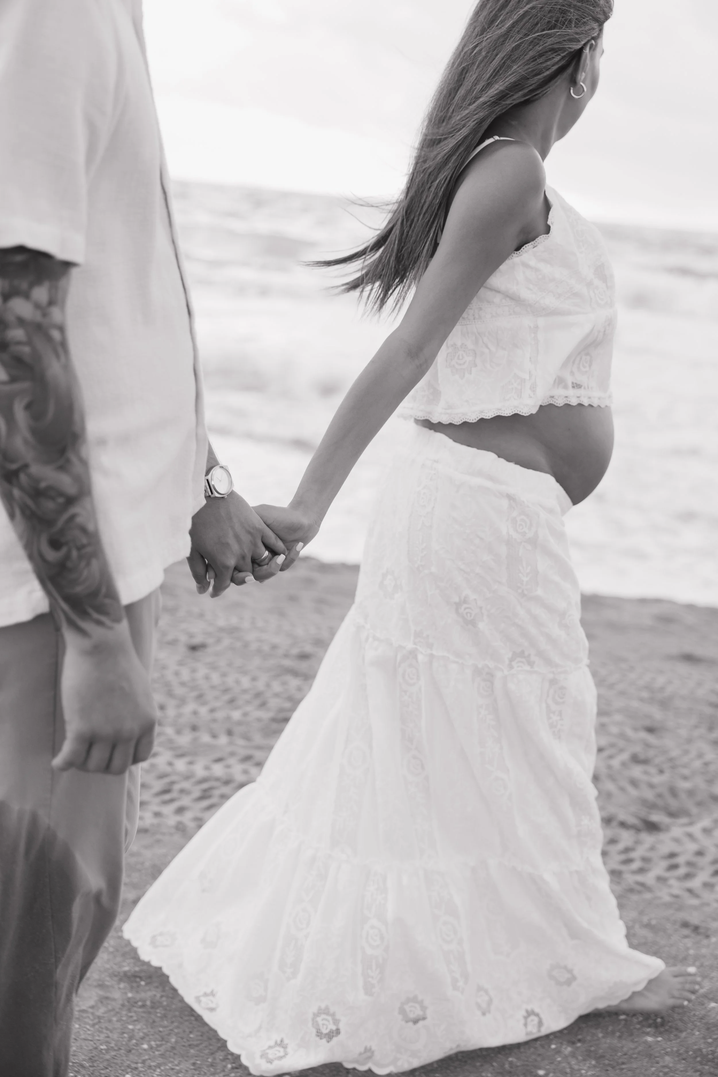 A pregnant woman in a flowing white dress holding hands with a man on the beach, with ocean waves in the background.