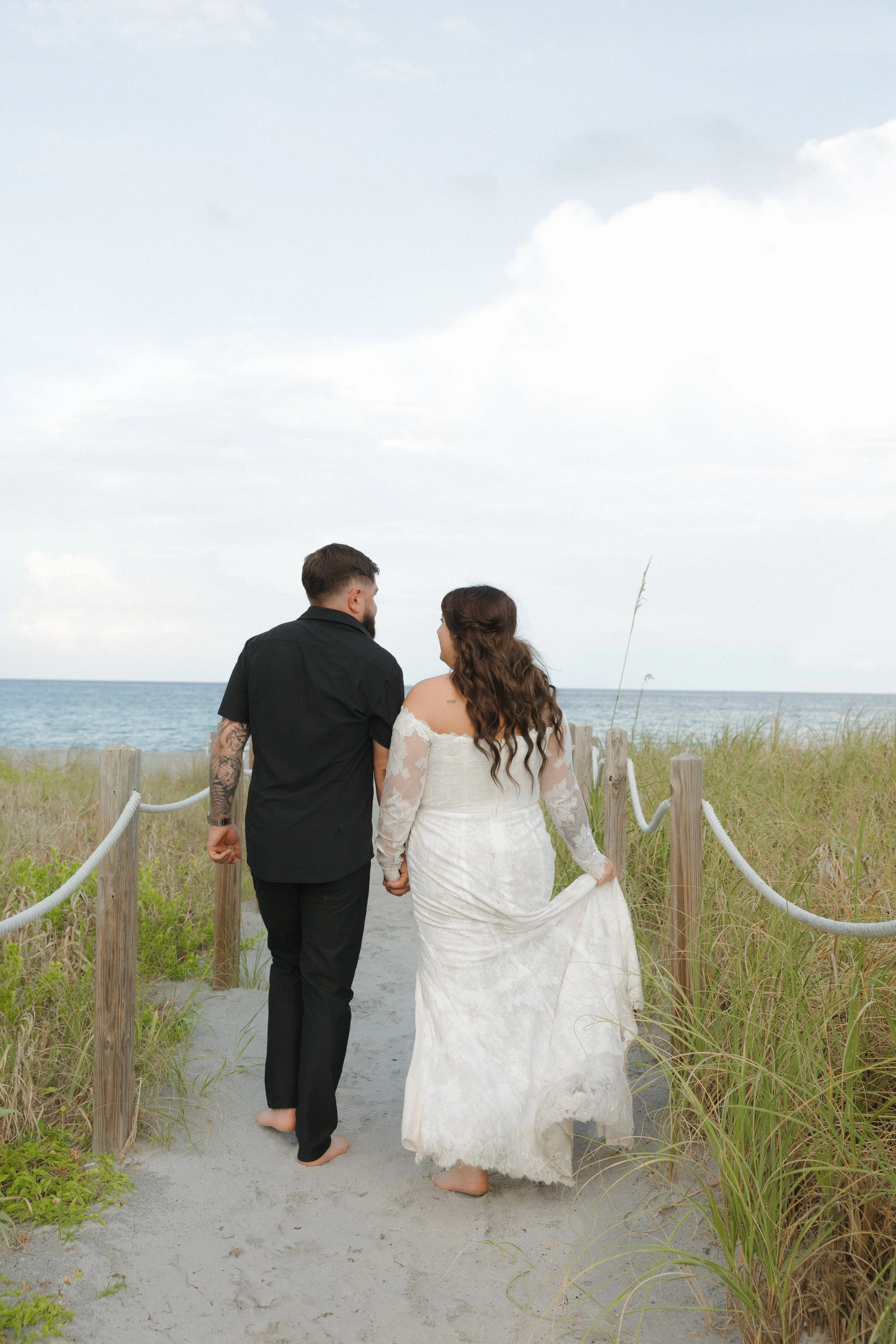 A couple holding hands walking barefoot on a sandy path toward the beach, with ocean and cloudy sky in the background.