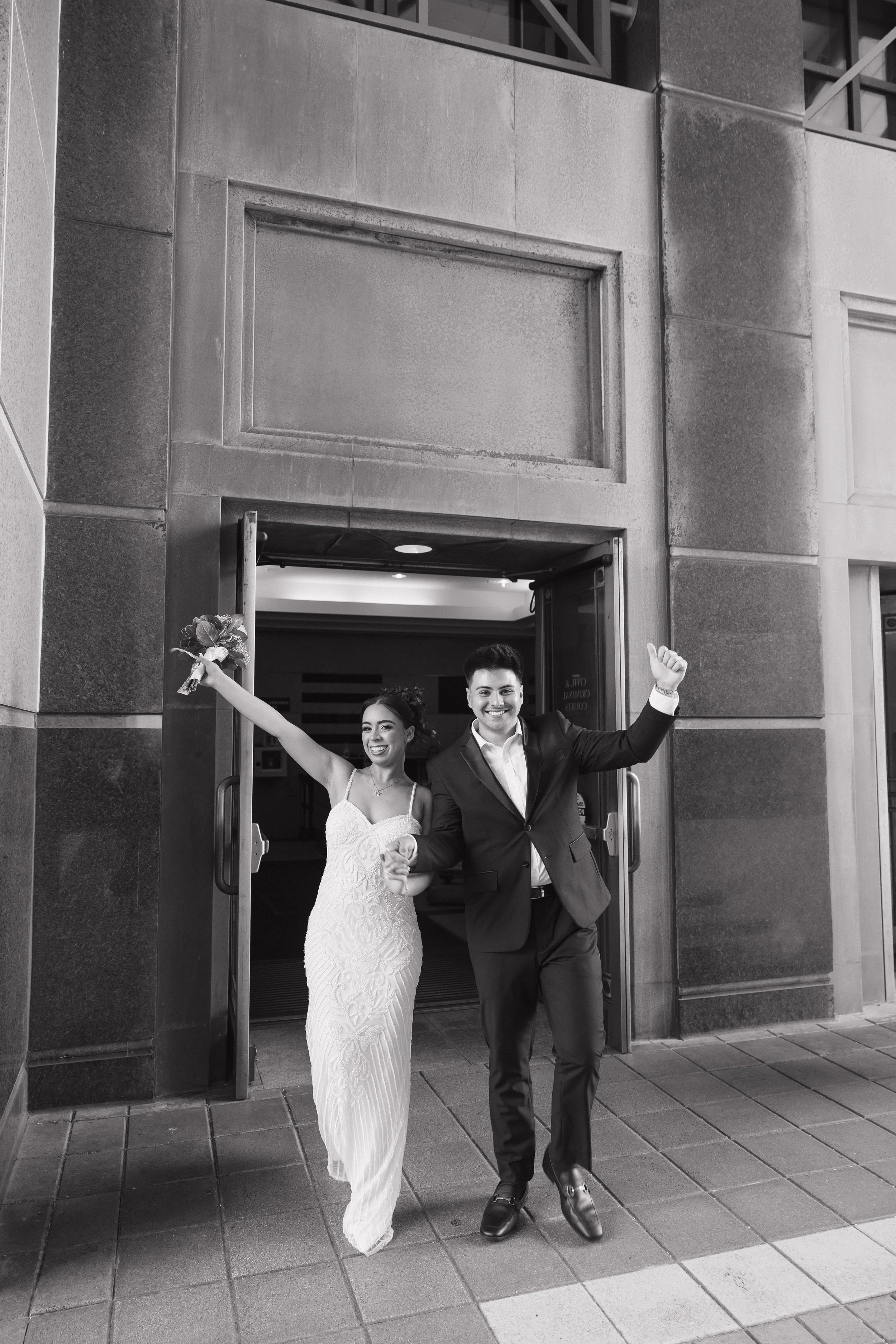 A newly married couple, a bride in a white wedding gown holding flowers and a groom in a tuxedo, cheerfully exiting a building through a doorway, celebrating their marriage.