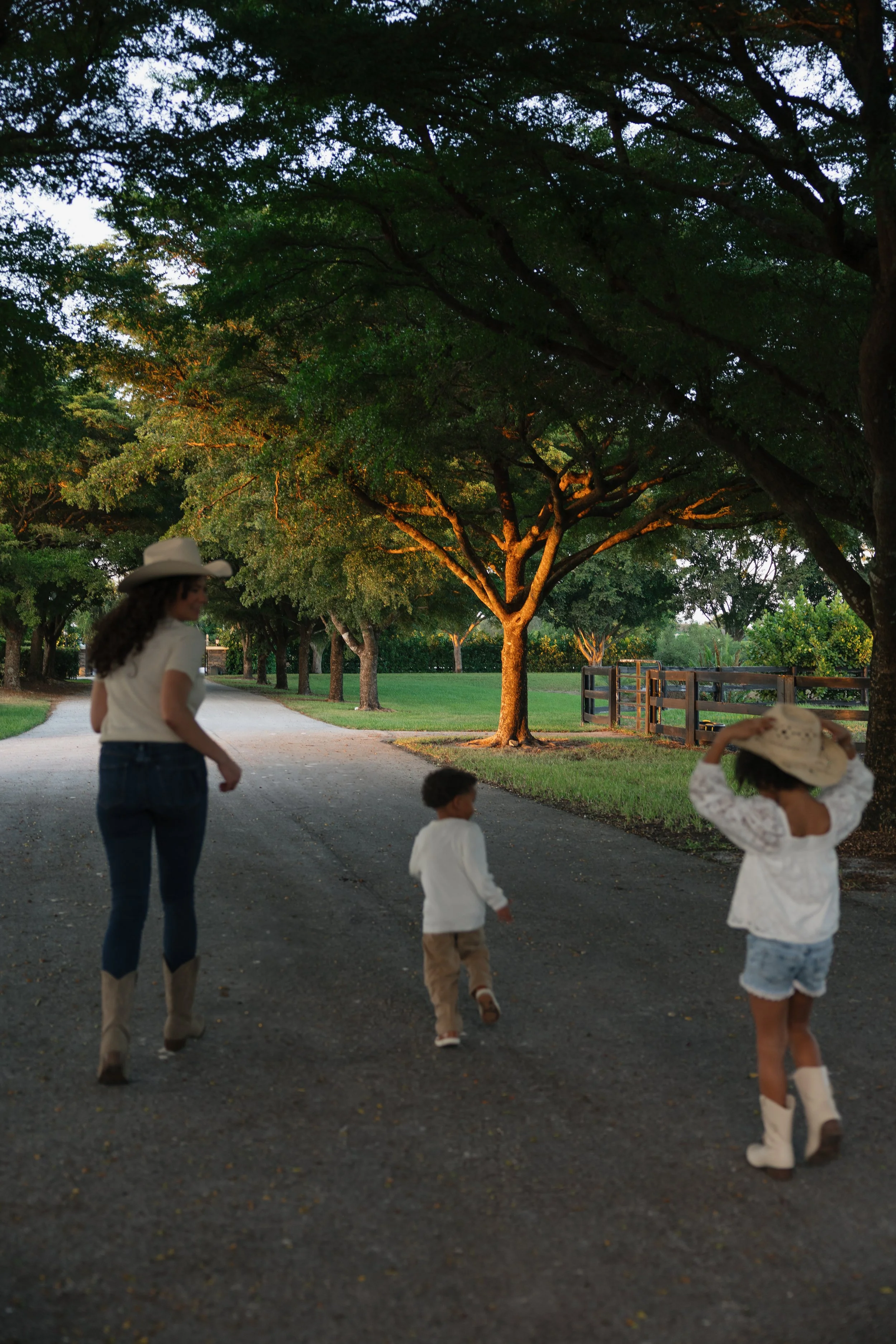 A woman and three children walking outdoors on a gravel path in a park, with trees and a wooden fence in the background during the evening.