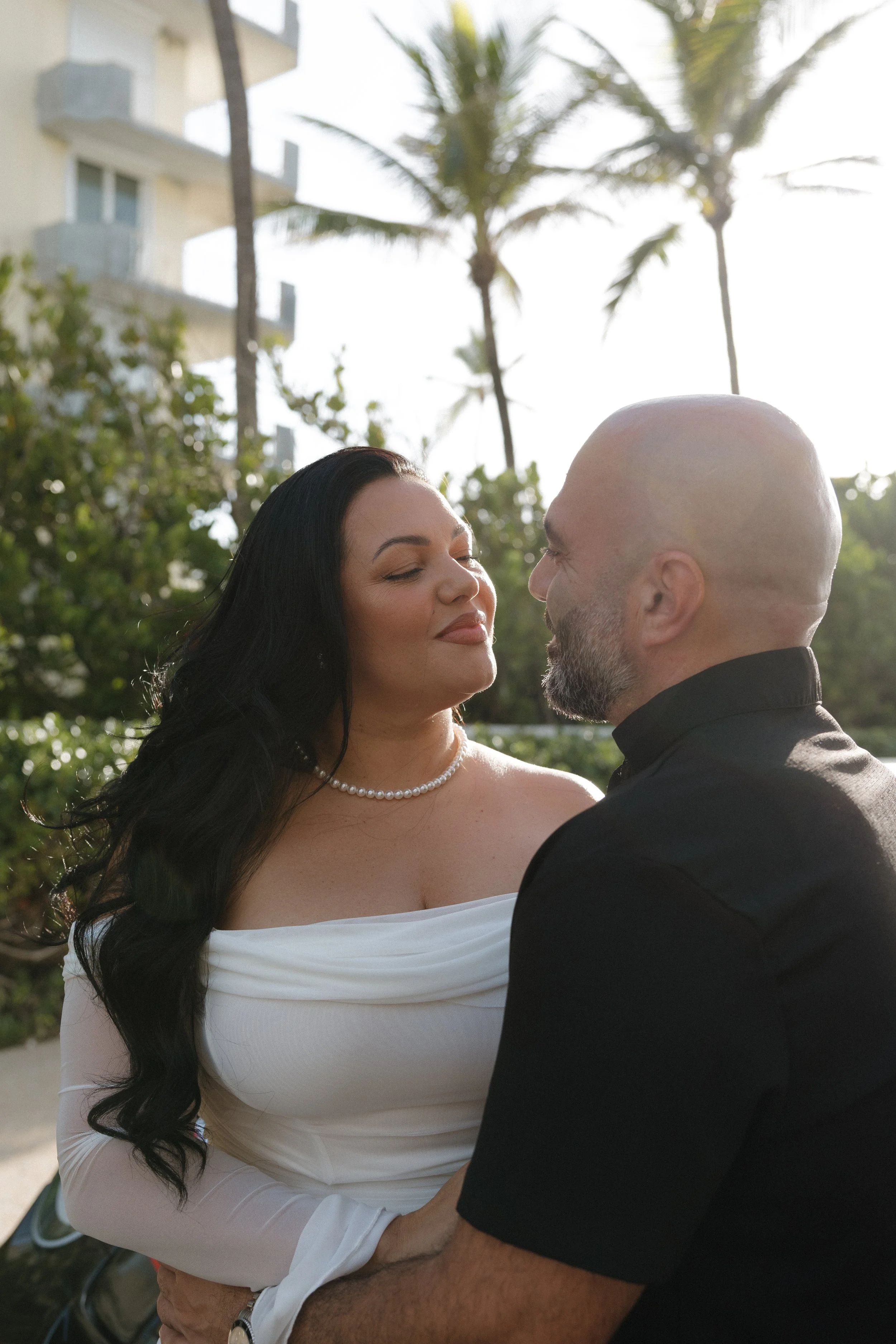 A couple standing close together outdoors, with the woman wearing a white off-shoulder top and pearl necklace, and the man in a black shirt, touching foreheads with their eyes closed. Palm trees and a building are in the background.