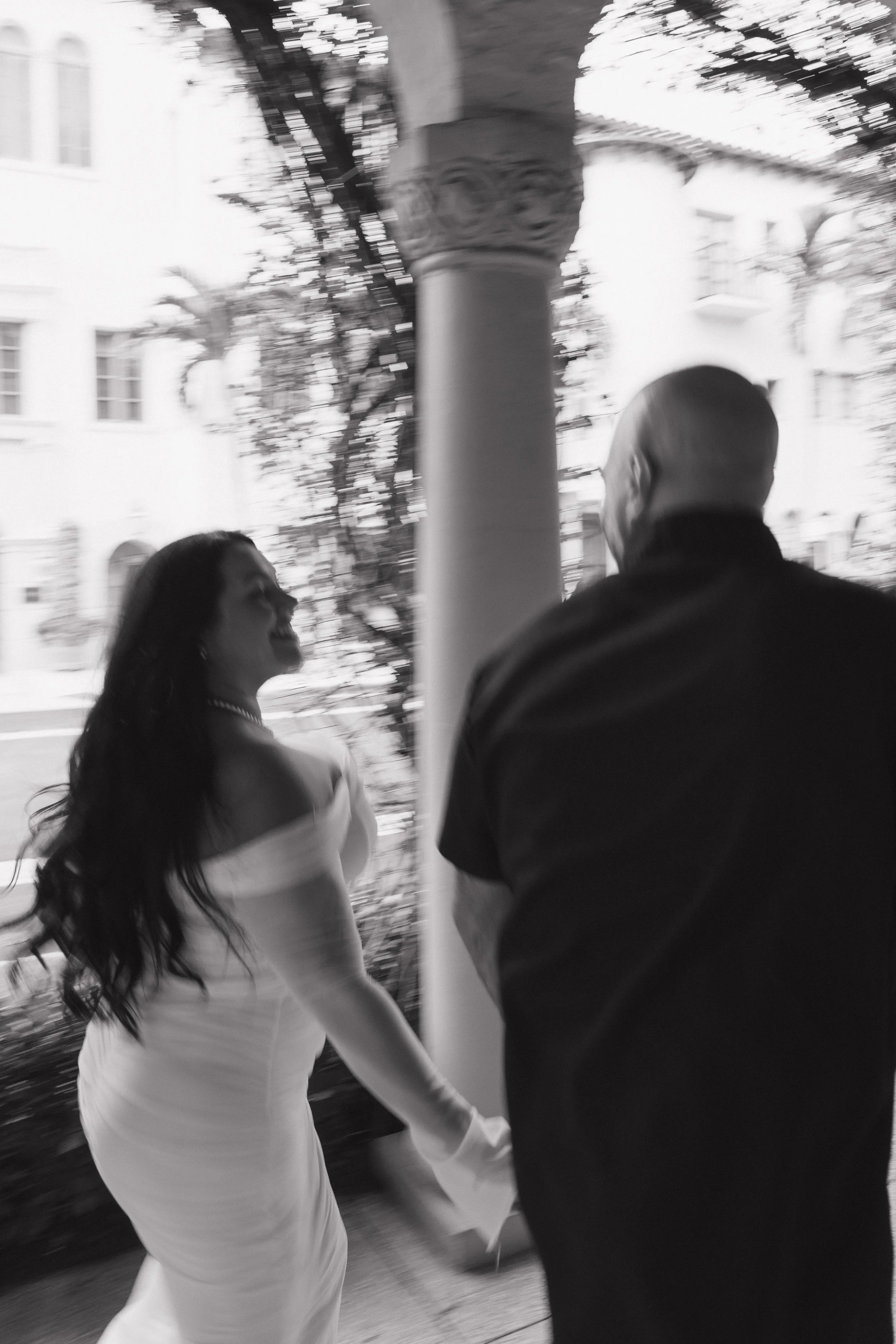 A black and white photo of a woman in a strapless dress smiling and holding hands with a man in a suit outdoors.