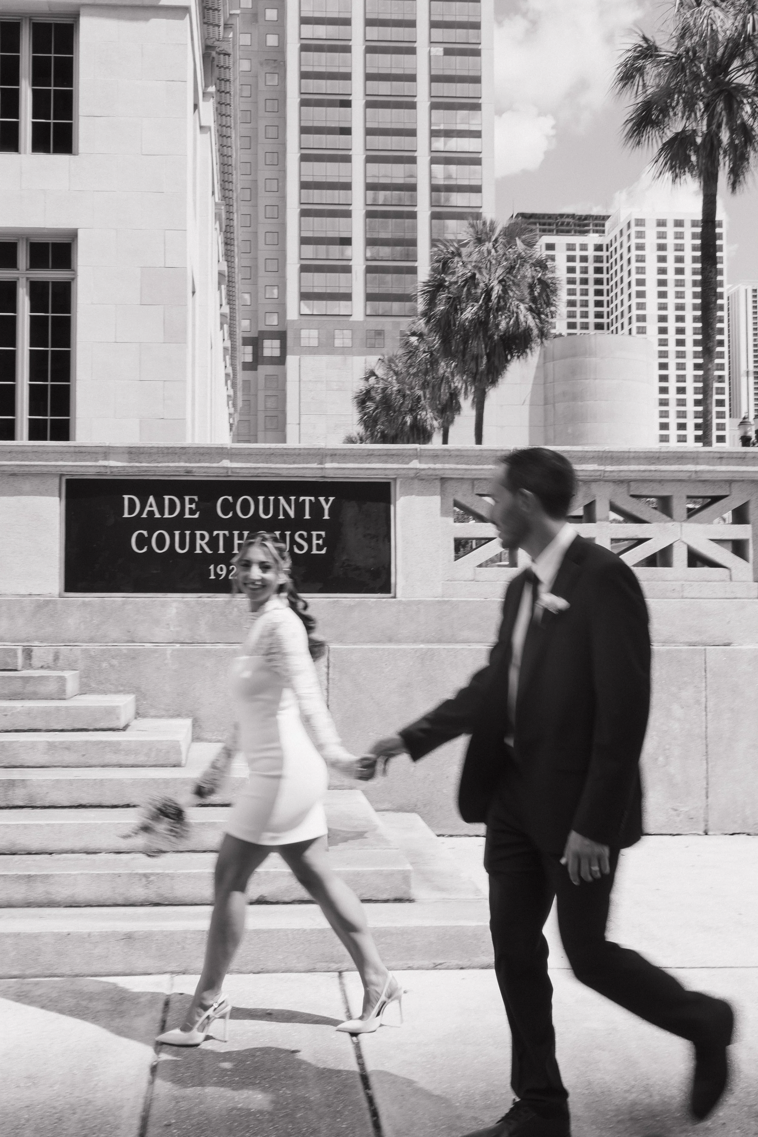 A woman in a white dress and heels holding hands with a man in a black suit walking down steps in front of a courthouse building with tall buildings and palm trees in the background.