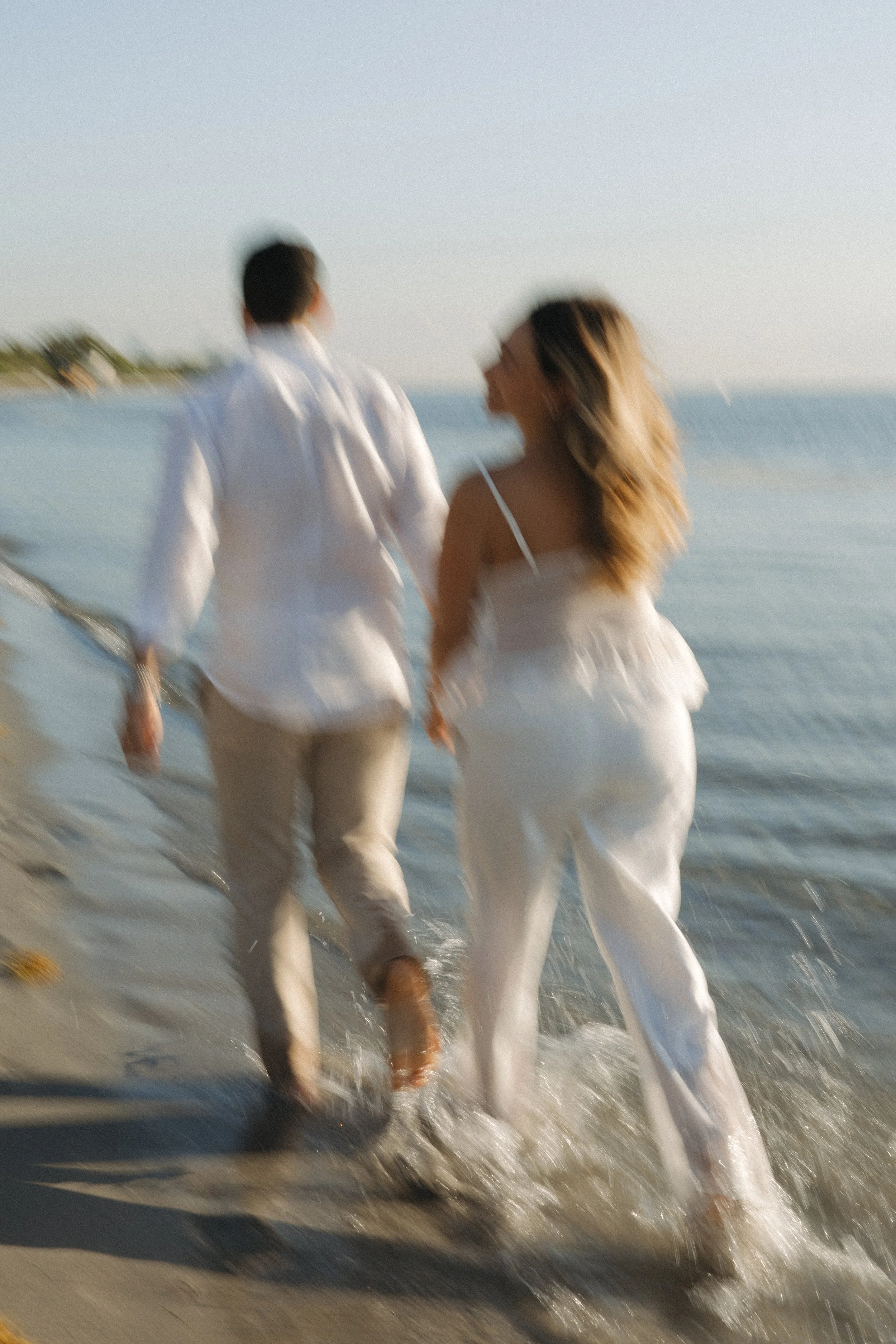 A couple walking on the beach holding hands, wearing white clothing, with ocean waves and a clear sky in the background.