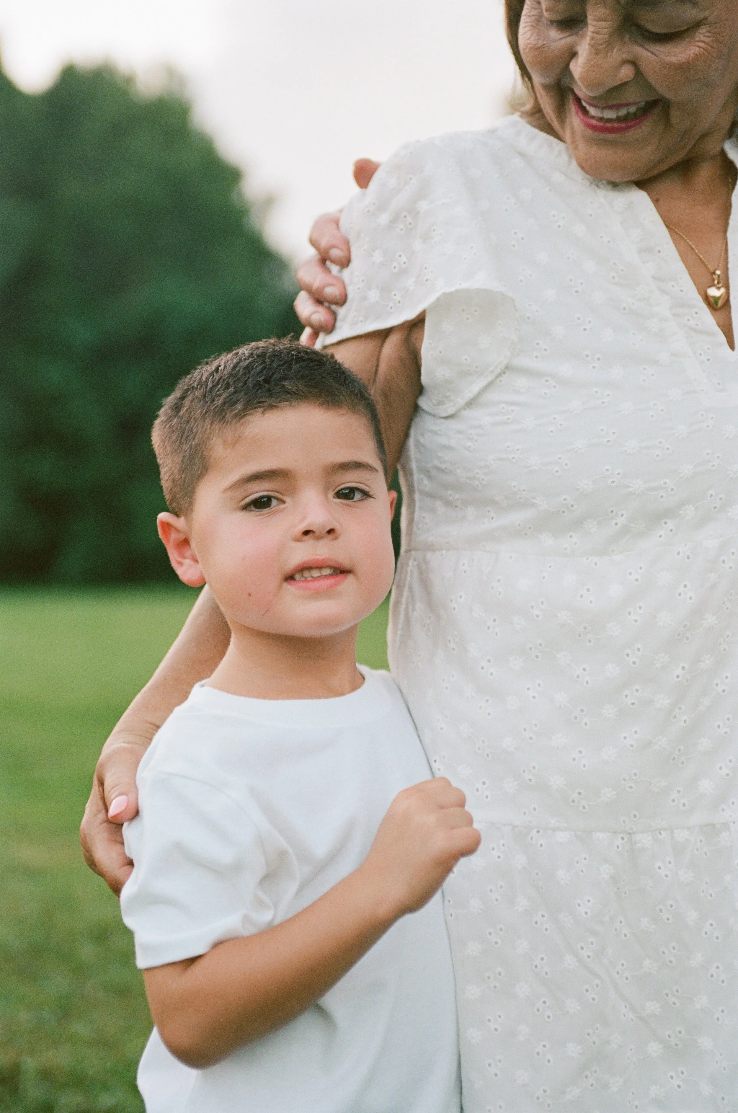 Close-up of an elderly woman and a young boy outdoors, smiling as they hold each other, with green trees in the background.