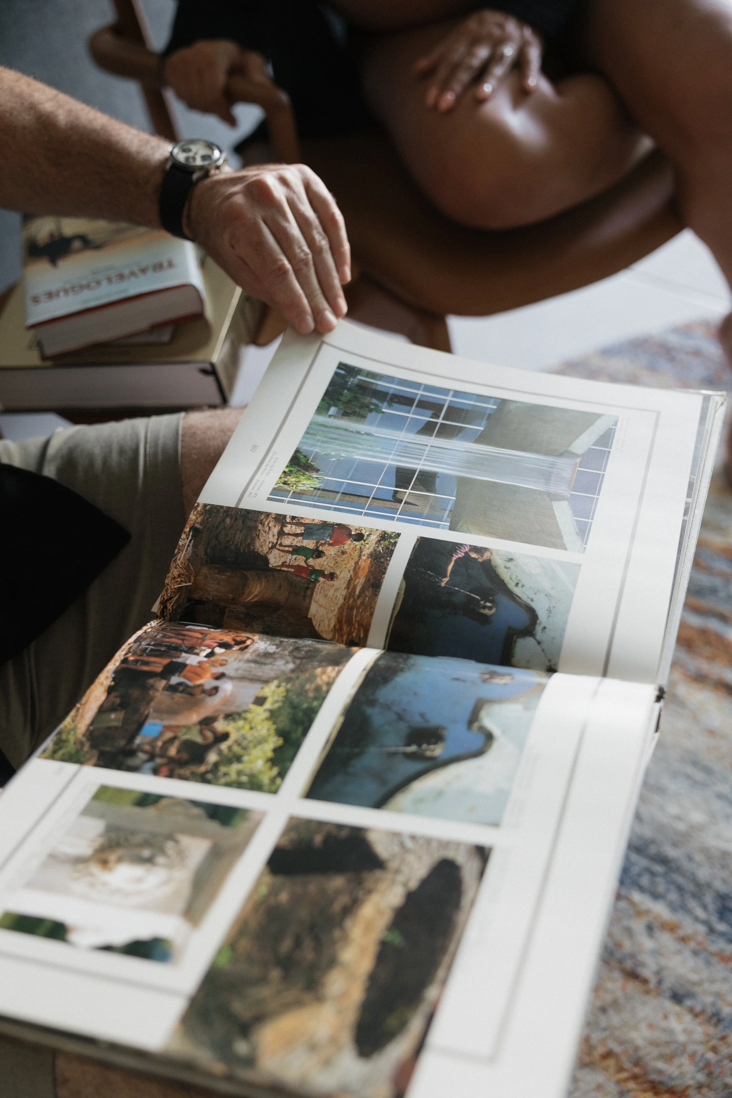 A person flipping through a magazine with colorful photographs of outdoor scenes, with a person sitting at a table in the background.