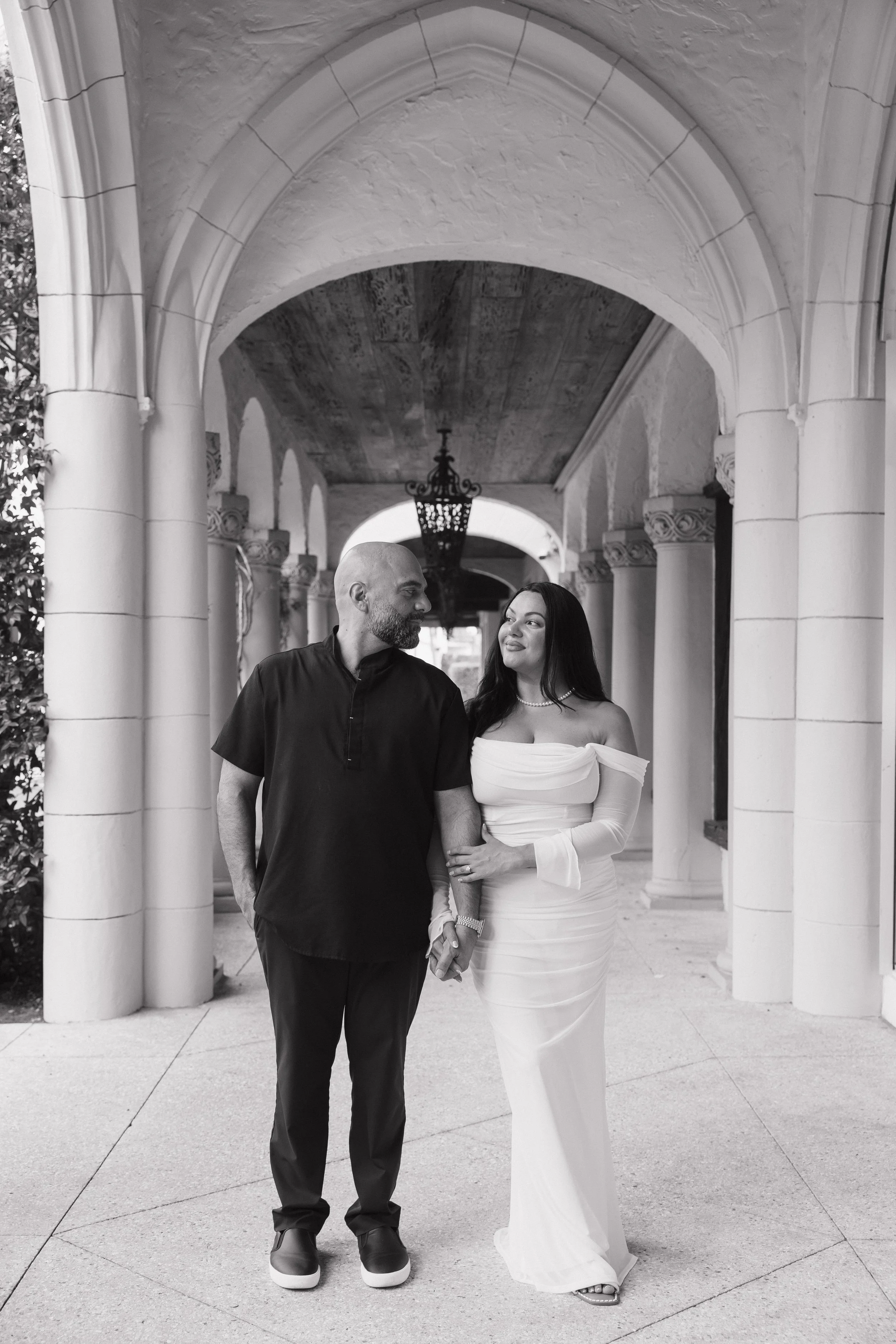 A man and woman holding hands under an arched entrance with columns, in a black and white photograph. The man is wearing a black shirt and dark pants, and the woman is in a white off-the-shoulder dress.