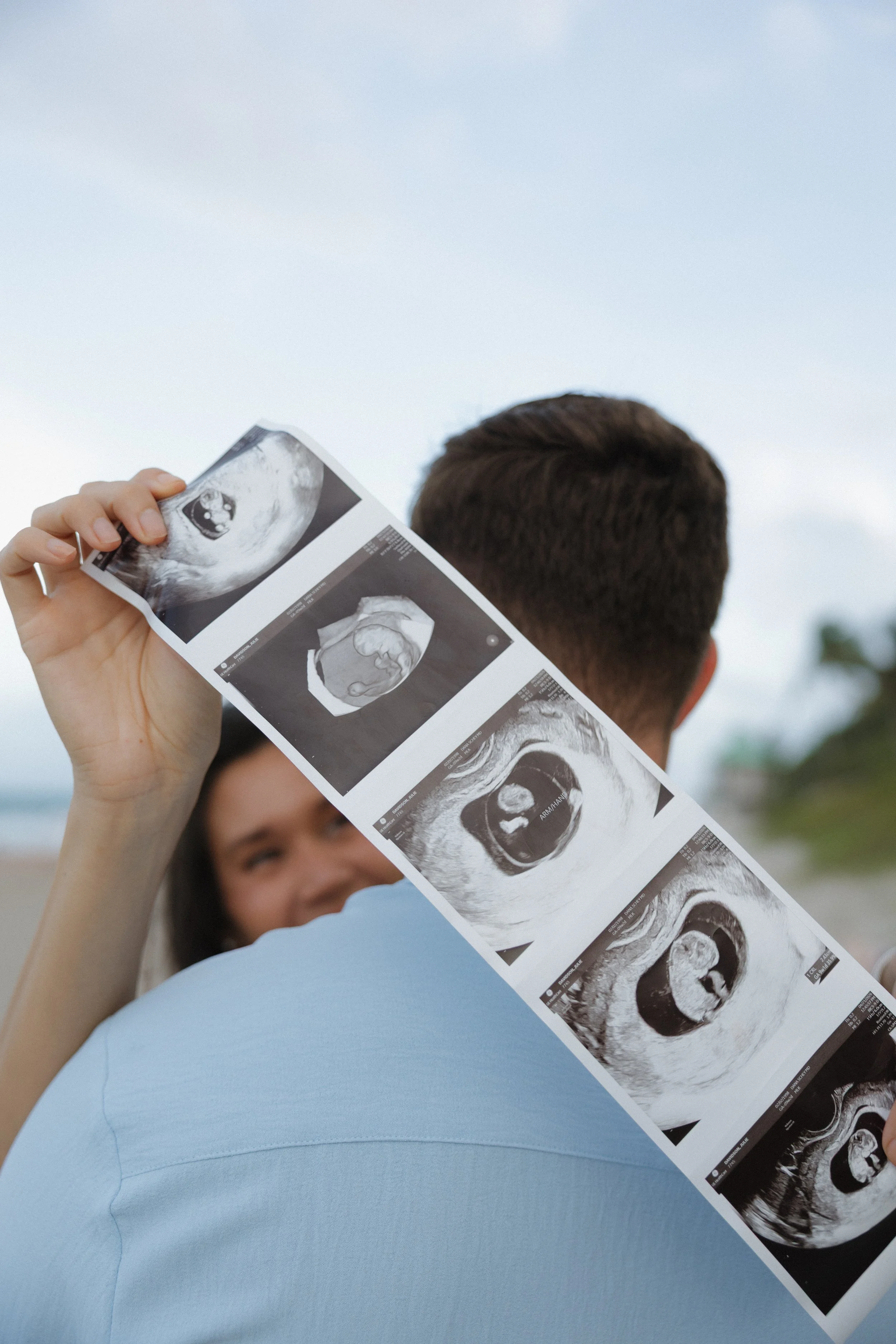 A person holding ultrasound images of a developing fetus, with part of their face and a young child's face visible in the background.