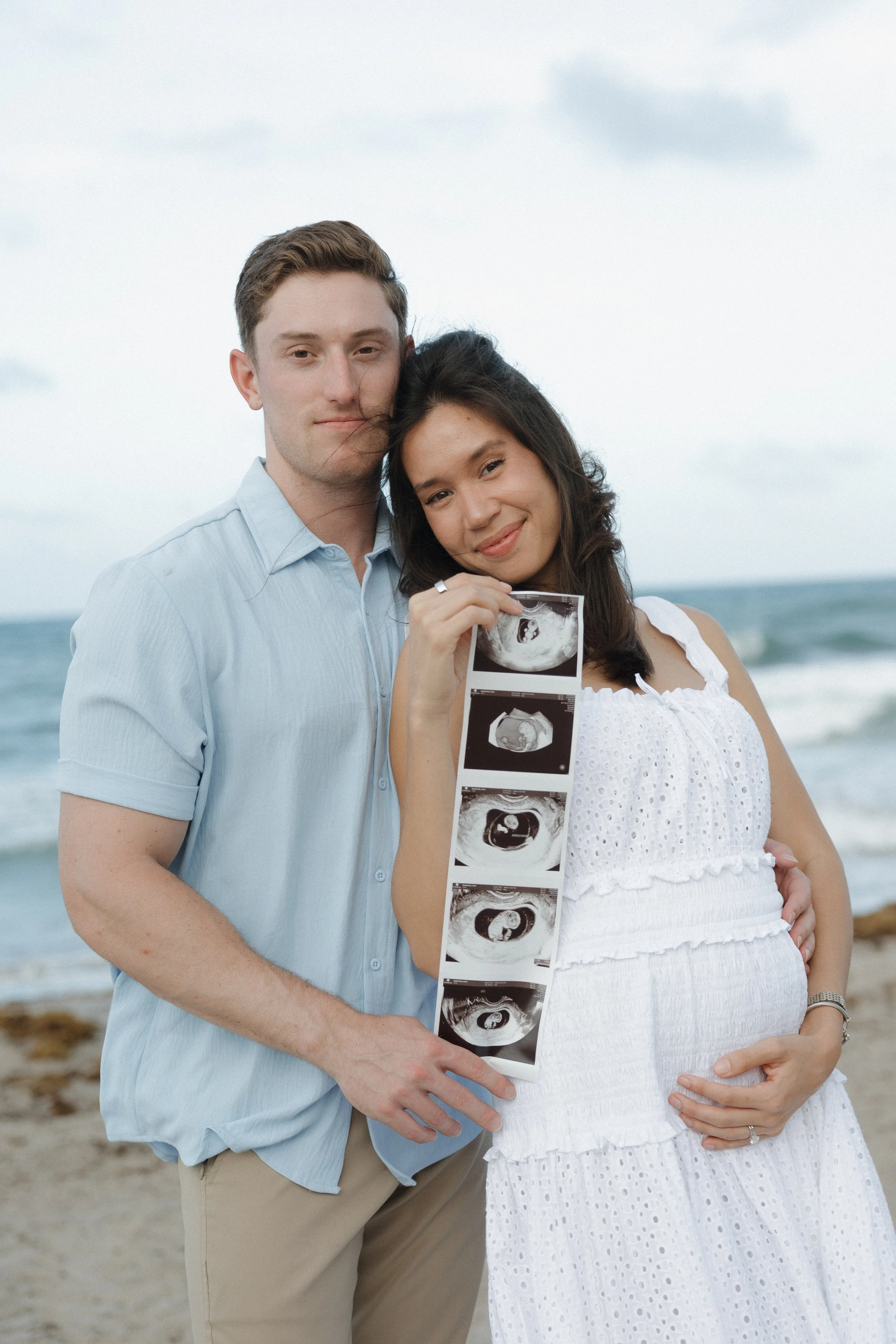 A couple at the beach during daytime, the woman is holding an ultrasound image and their pregnancy is visible, with the woman wearing a white dress and the man in a light blue shirt.
