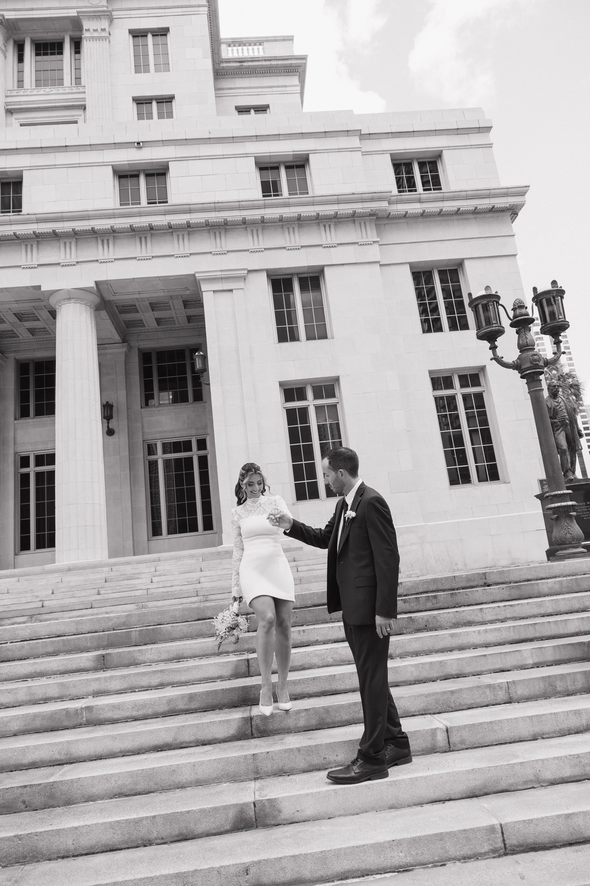 A black and white photograph of a couple on the steps of a large, classical-style government building with columns and multiple windows. The woman is wearing a white dress and heels, holding a bouquet, and the man is dressed in a dark suit, holding h
