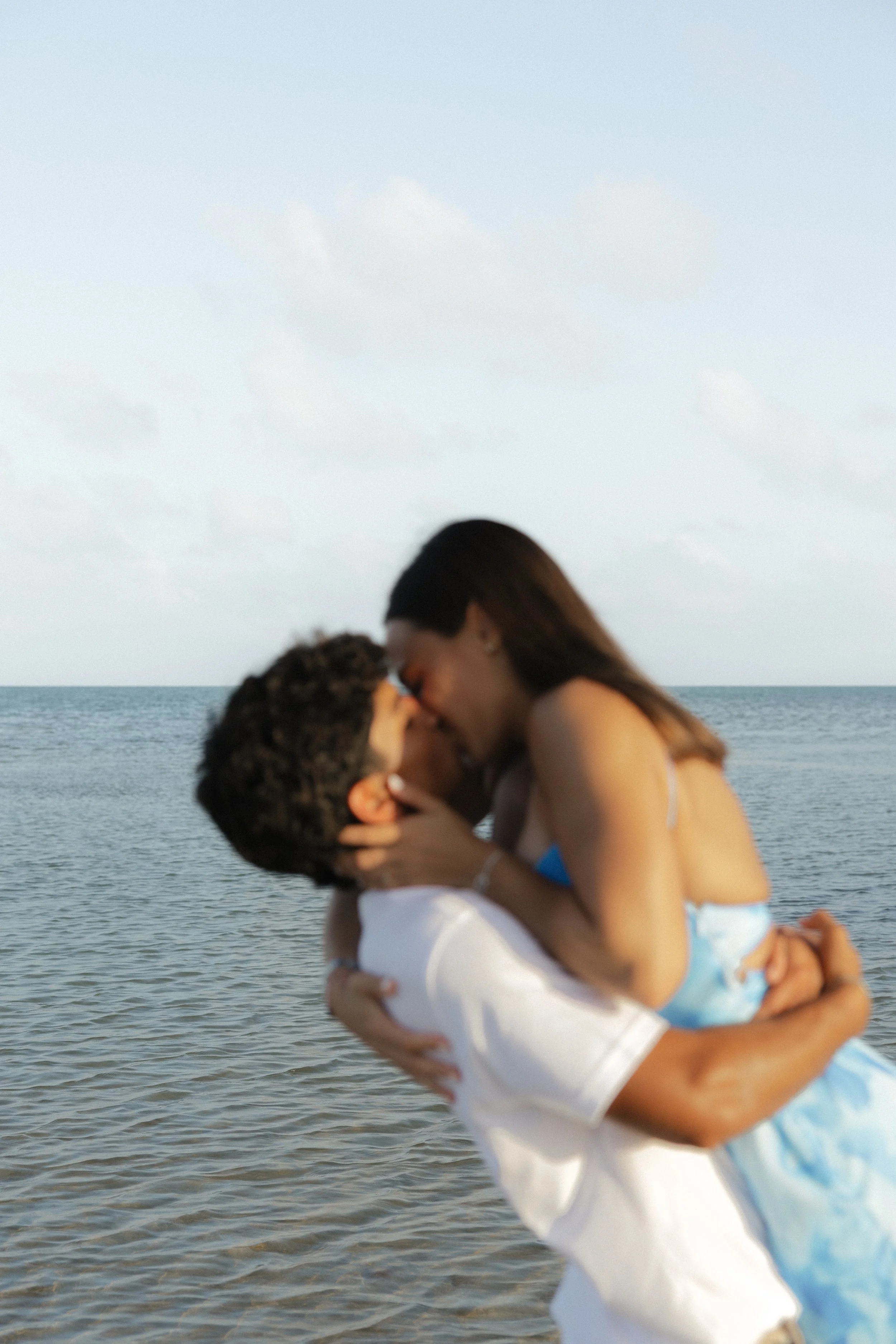 A couple sharing a kiss on the beach, with the ocean and sky in the background.