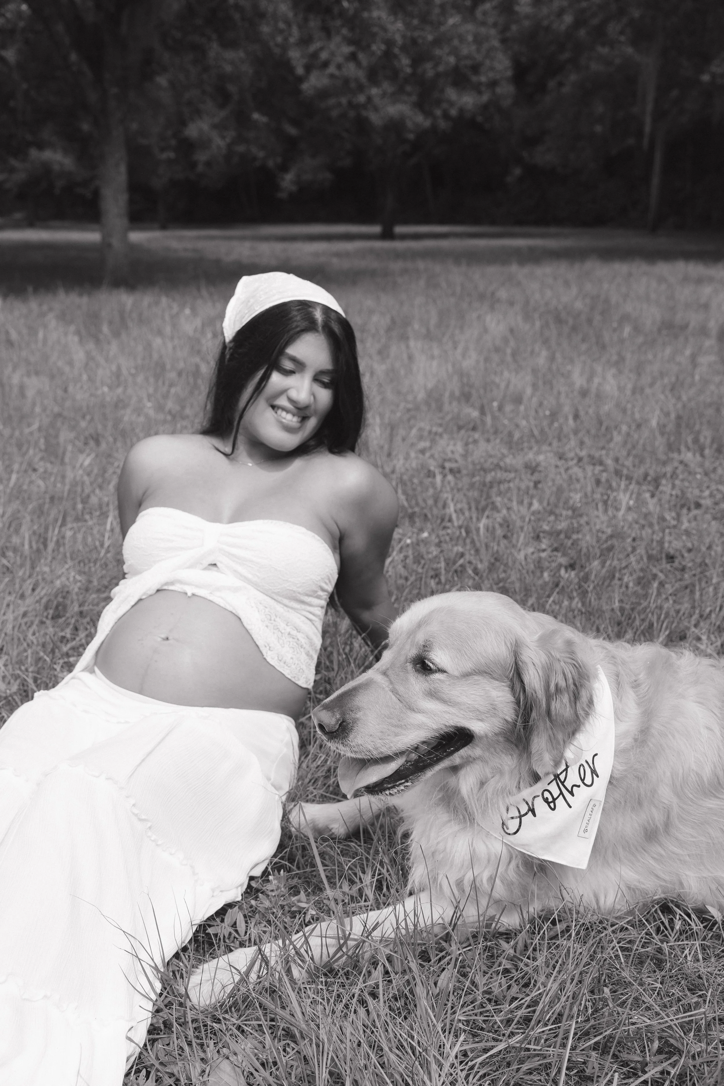 A woman smiling and sitting in a grassy field next to a large golden retriever dog wearing a bandana. The woman is wearing a white head covering and a strapless top. The scene is outdoors with trees in the background, and the photo is in black and wh