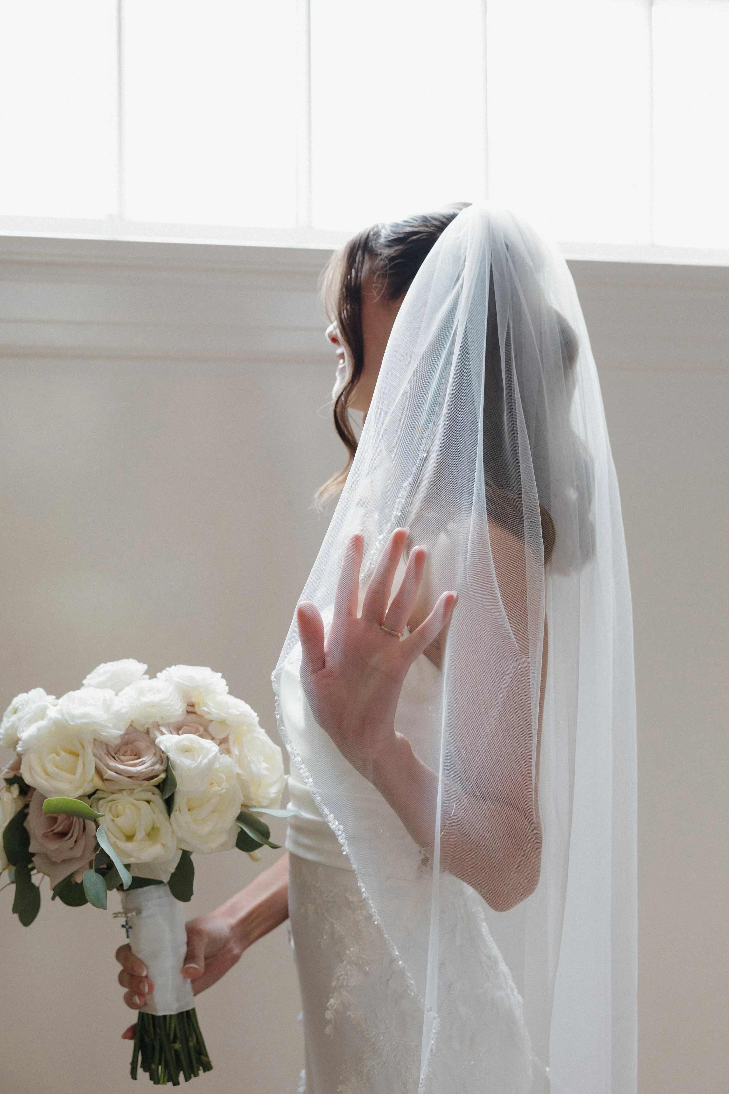 A bride in a white wedding dress with lace details, wearing a sheer veil, is holding a bouquet of white and blush roses with green leaves, standing near a window with natural light.