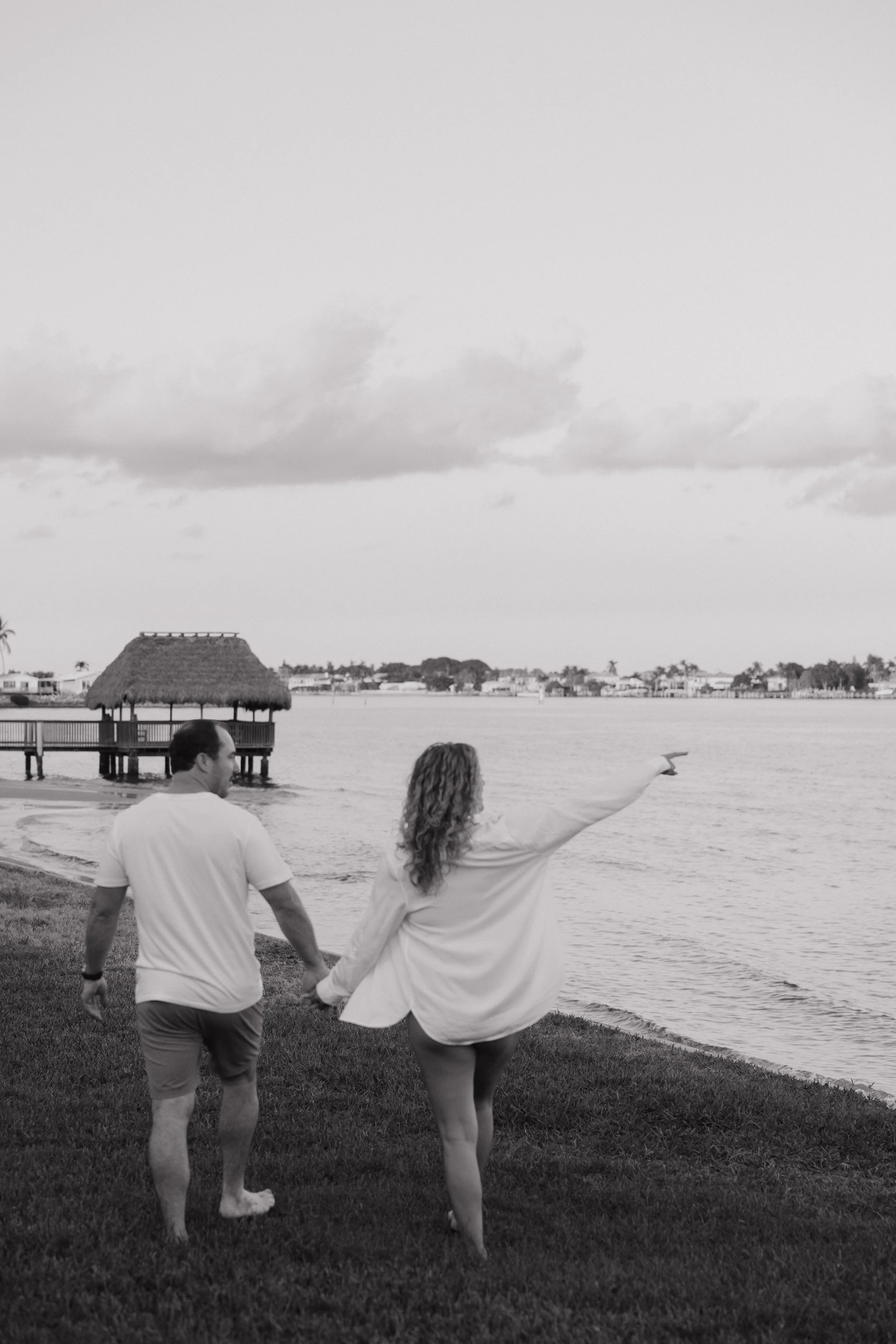A man and a woman holding hands and walking along the grassy shore of a body of water, with a thatched gazebo on stilts over the water, in front of a distant shoreline with houses and trees, under a cloudy sky.