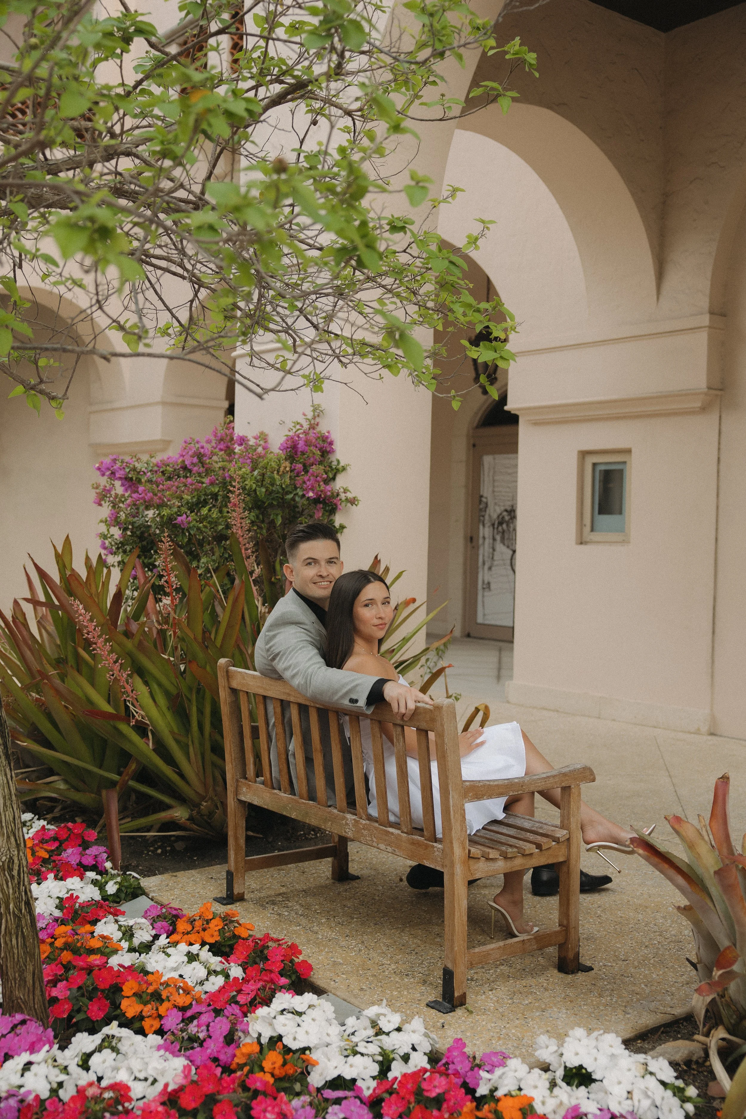 A man and woman sitting on a wooden bench in a garden, surrounded by colorful flowers and green plants, with arched building structures in the background.