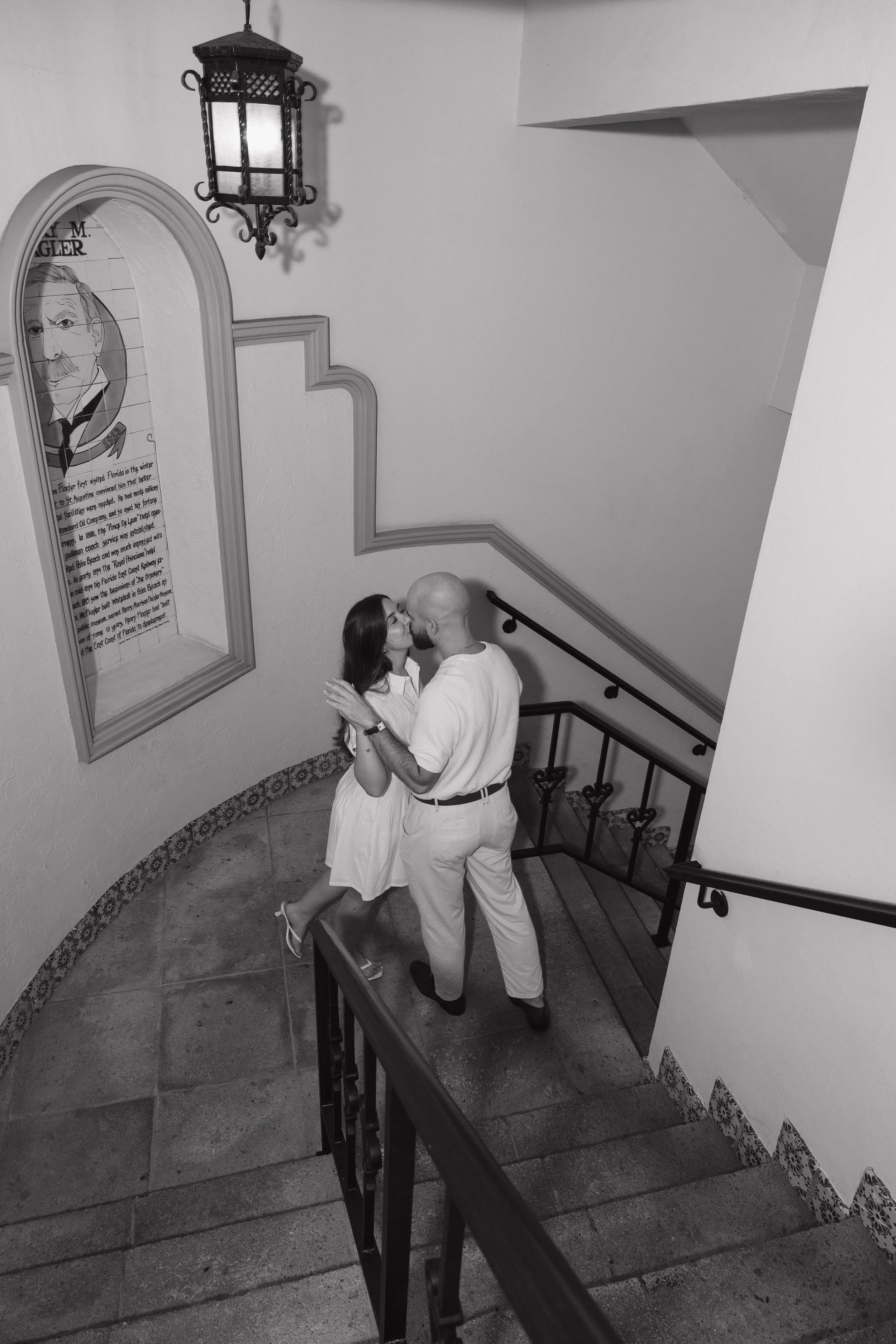 A couple sharing a kiss on a staircase landing, surrounded by white walls with decorative trim, a large framed portrait, and a hanging lantern-style light fixture.