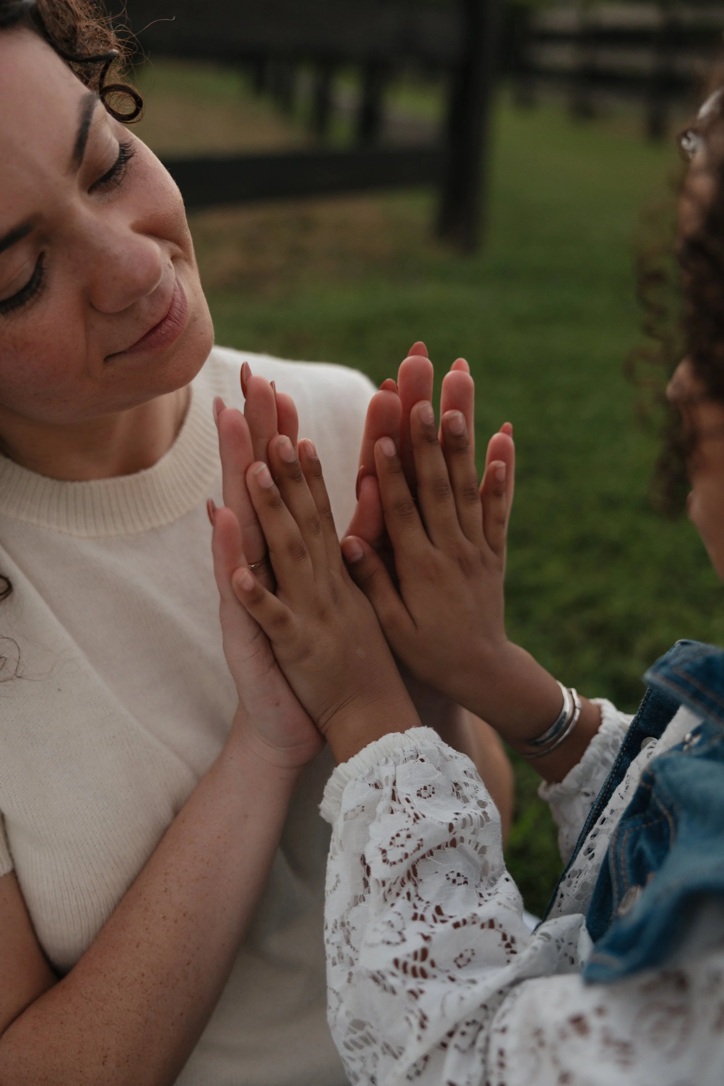 A woman and a young girl pressing their palms together in a gesture of connection outdoors with a wooden fence and green grass in the background.