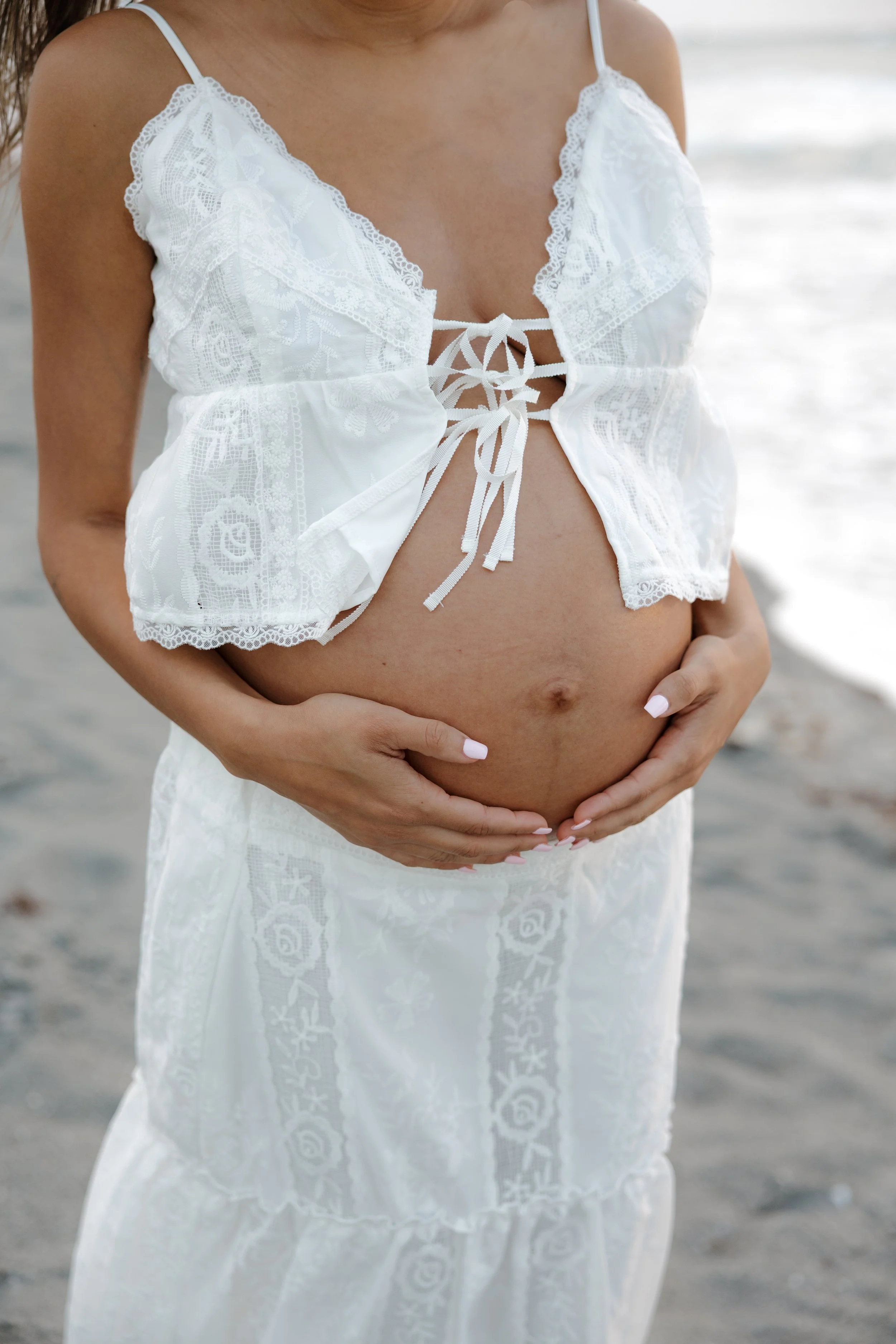 A pregnant woman in a white lace dress is holding her belly at the beach.