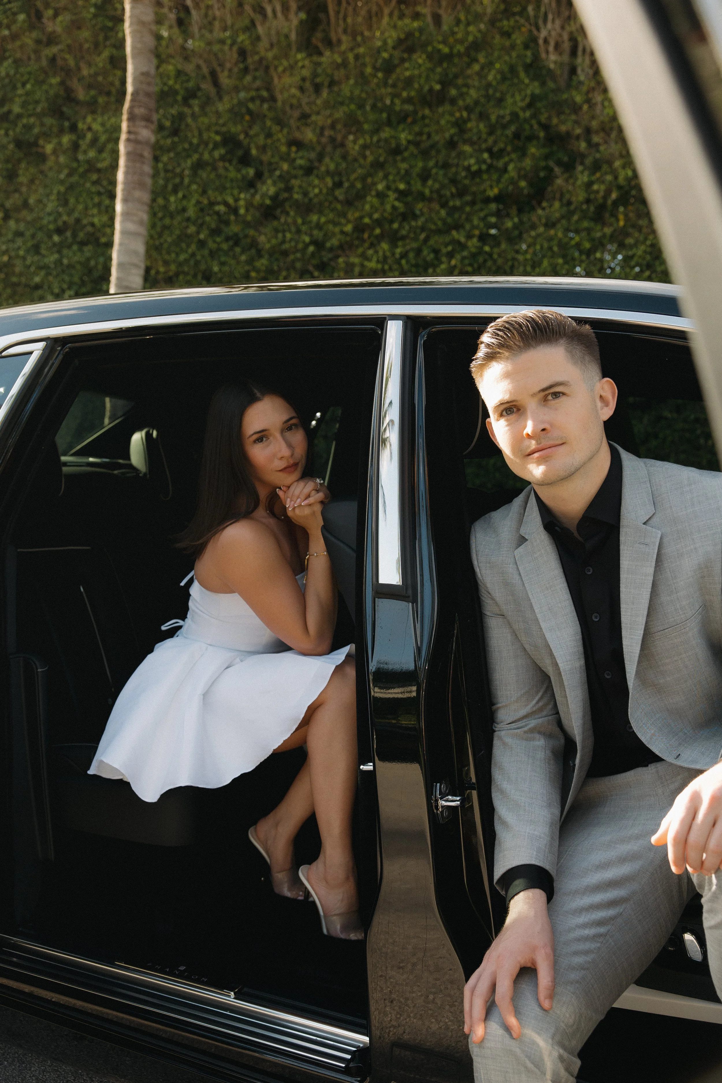 A man and woman in formal attire next to a black car, with the woman sitting inside and the man leaning out of the car window.